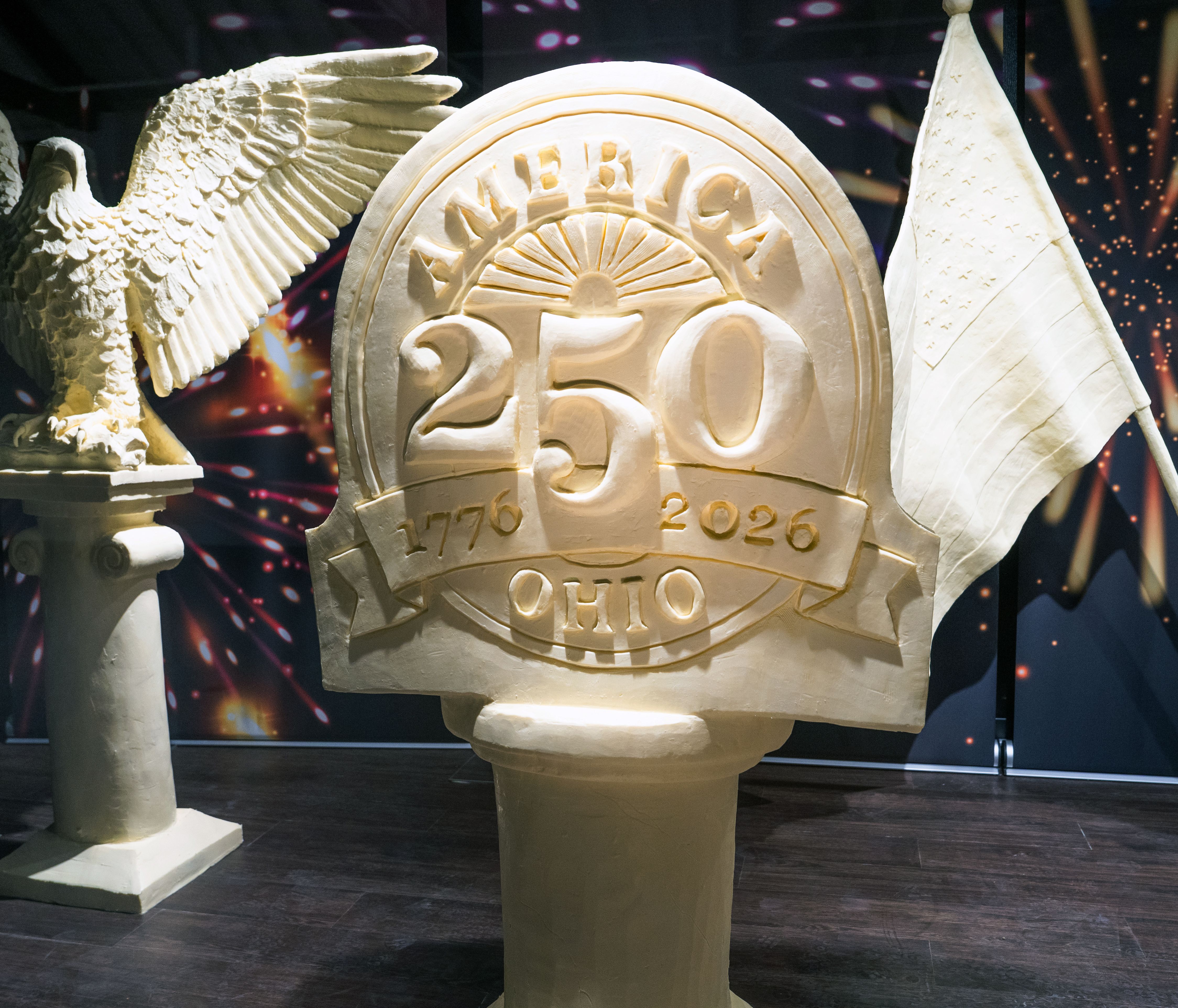 A sign that reads "America 250 Ohio - 1776-2026" sculpted entirely of butter, next to a butter bald eagle and American flag, amid a backdrop of fireworks.