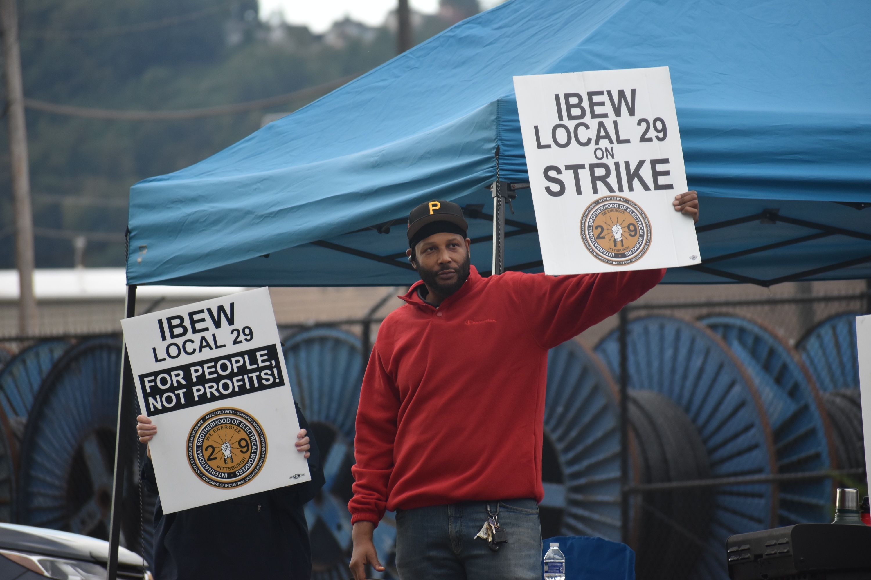 Protester in a red shirt holds a sign that reads "IBEW LOCAL 29 ON STRIKE" beside another sign; blue tent backdrop and a circular union emblem on the signs.