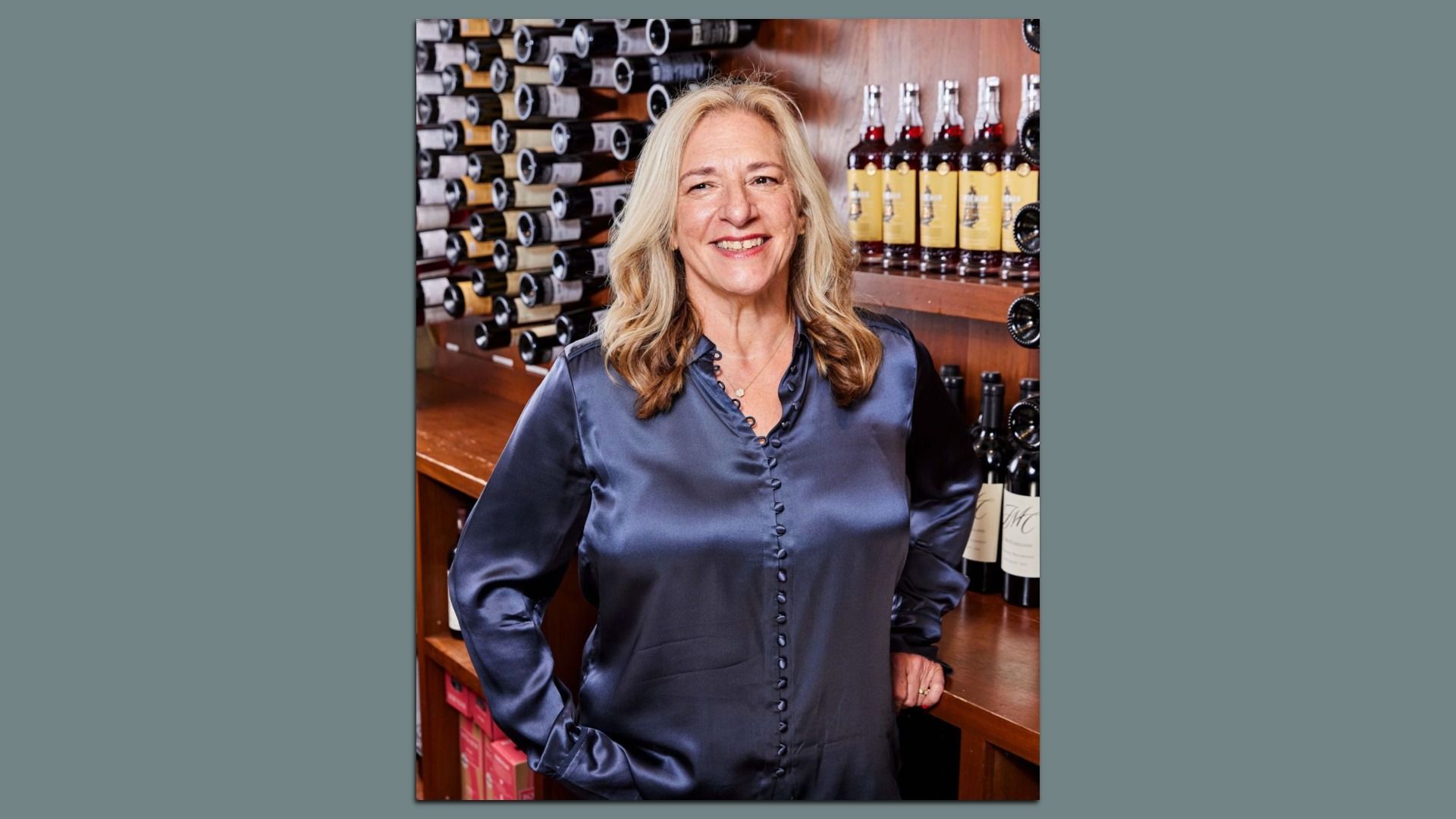 Smiling middle-aged woman with blonde hair in a blue satin blouse stands in a wine shop, with shelves of bottles behind her.