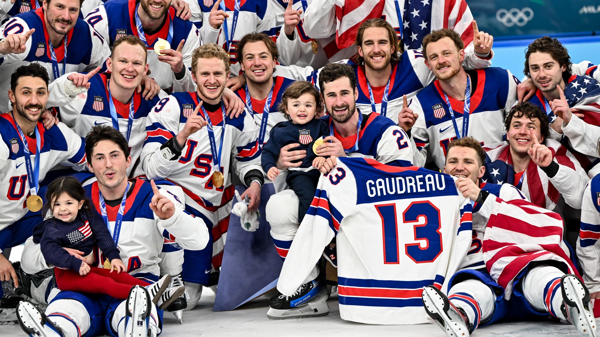 The USA men's ice hockey team celebrates winning gold on the ice, posing for a photo with Johnny Gaudreau's kids and jersey.