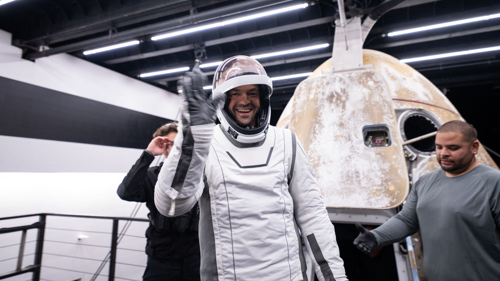 Photo of a man smiling and waving while emerging from a space capsule in a spacesuit.