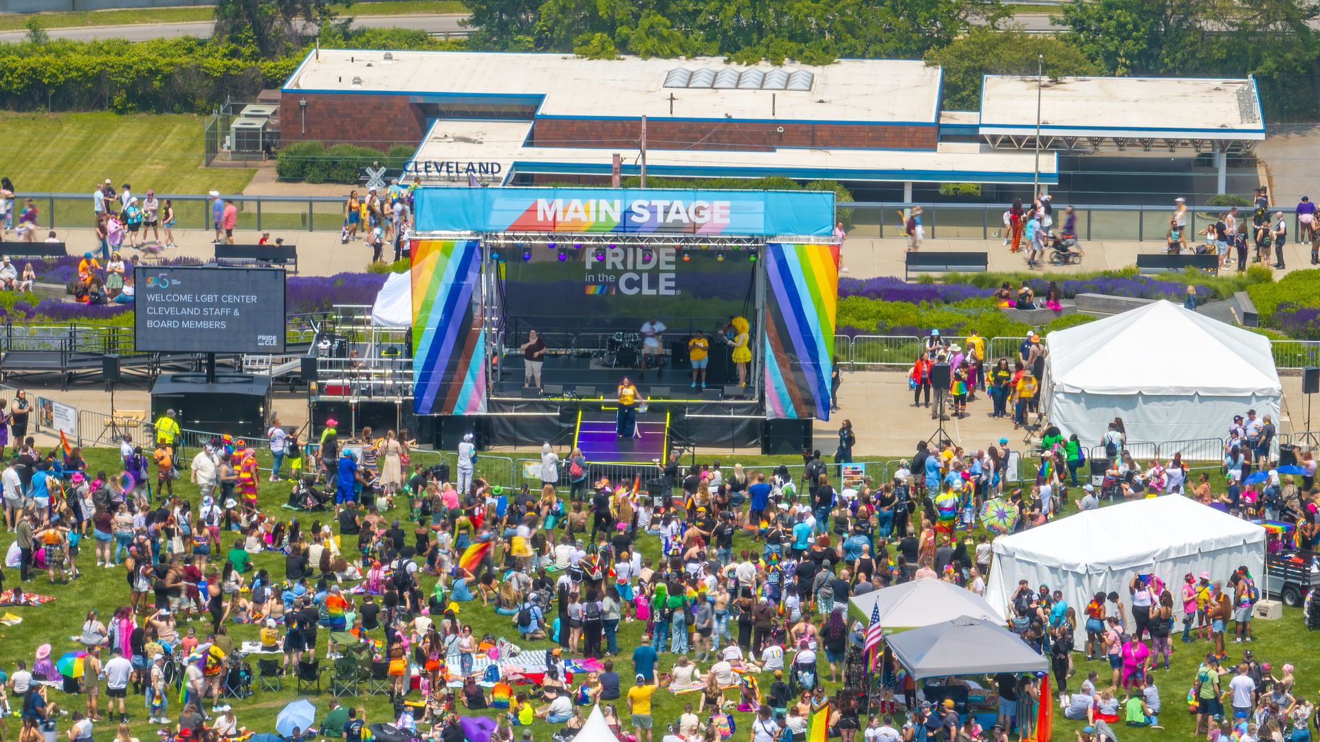 Outdoor Pride festival with a rainbow-decorated main stage and a grassy crowd; a large left screen reads "WELCOME LGBT CENTER" as people walk, sit, and gather around colorful tents.