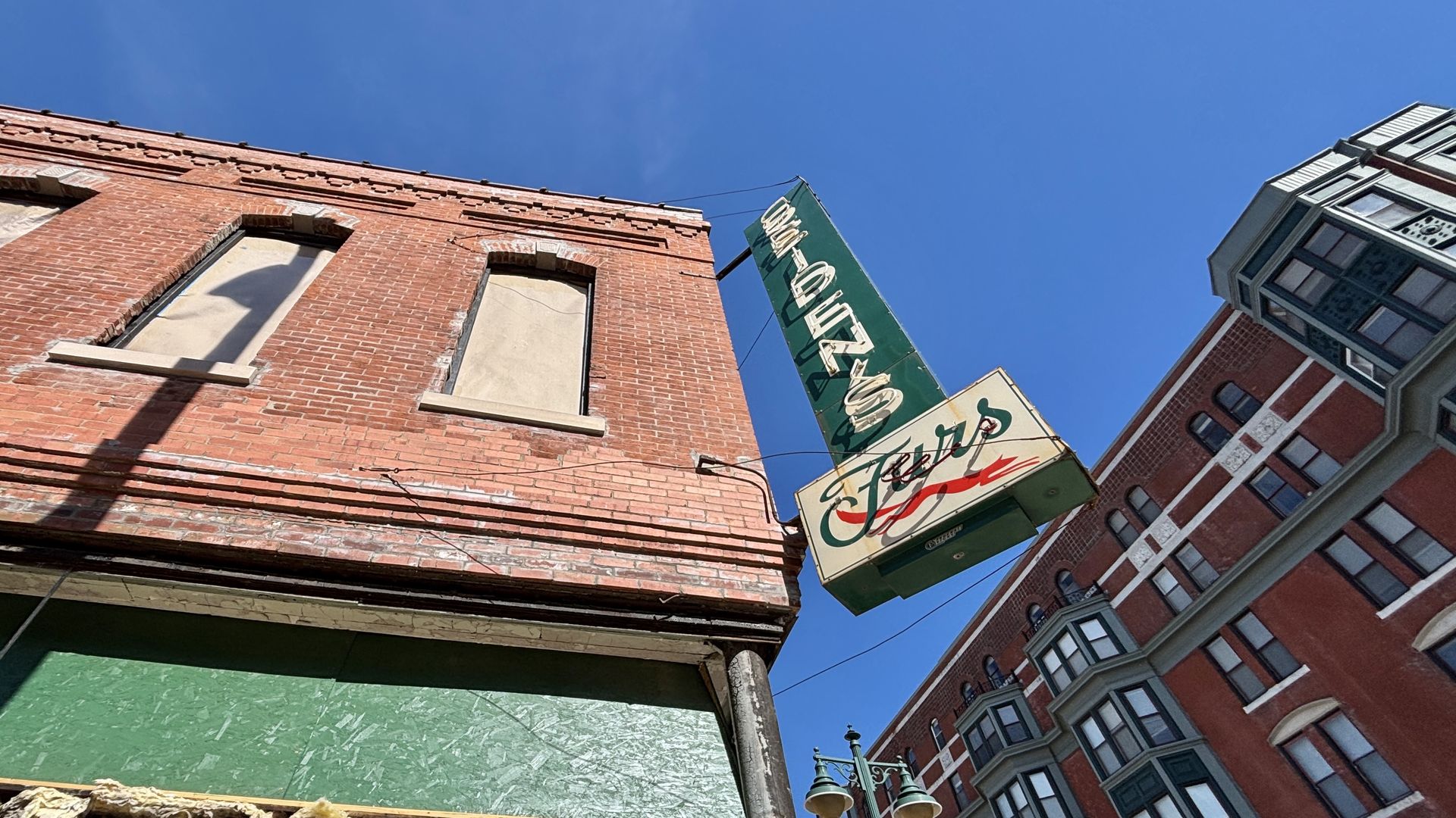 Blue sky over a brick storefront with boarded windows. A green hanging sign with white lettering reading "SEIDEN'S" extends diagonally from the building; a neighboring brick building with bay windows rises on the right.