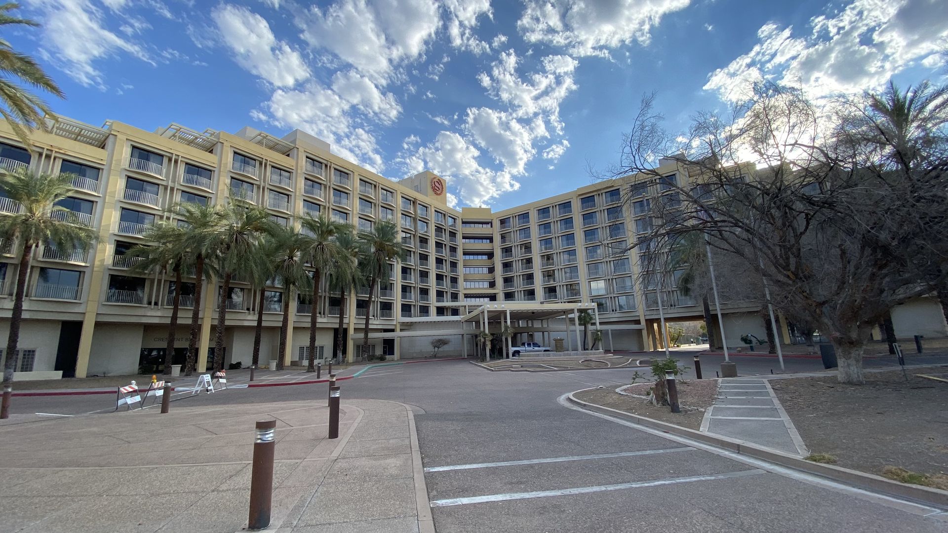Wide view of a beige, multi-story hotel with repeating balconies, palm trees along the front, a covered drop-off, and a bright blue sky with scattered white clouds.