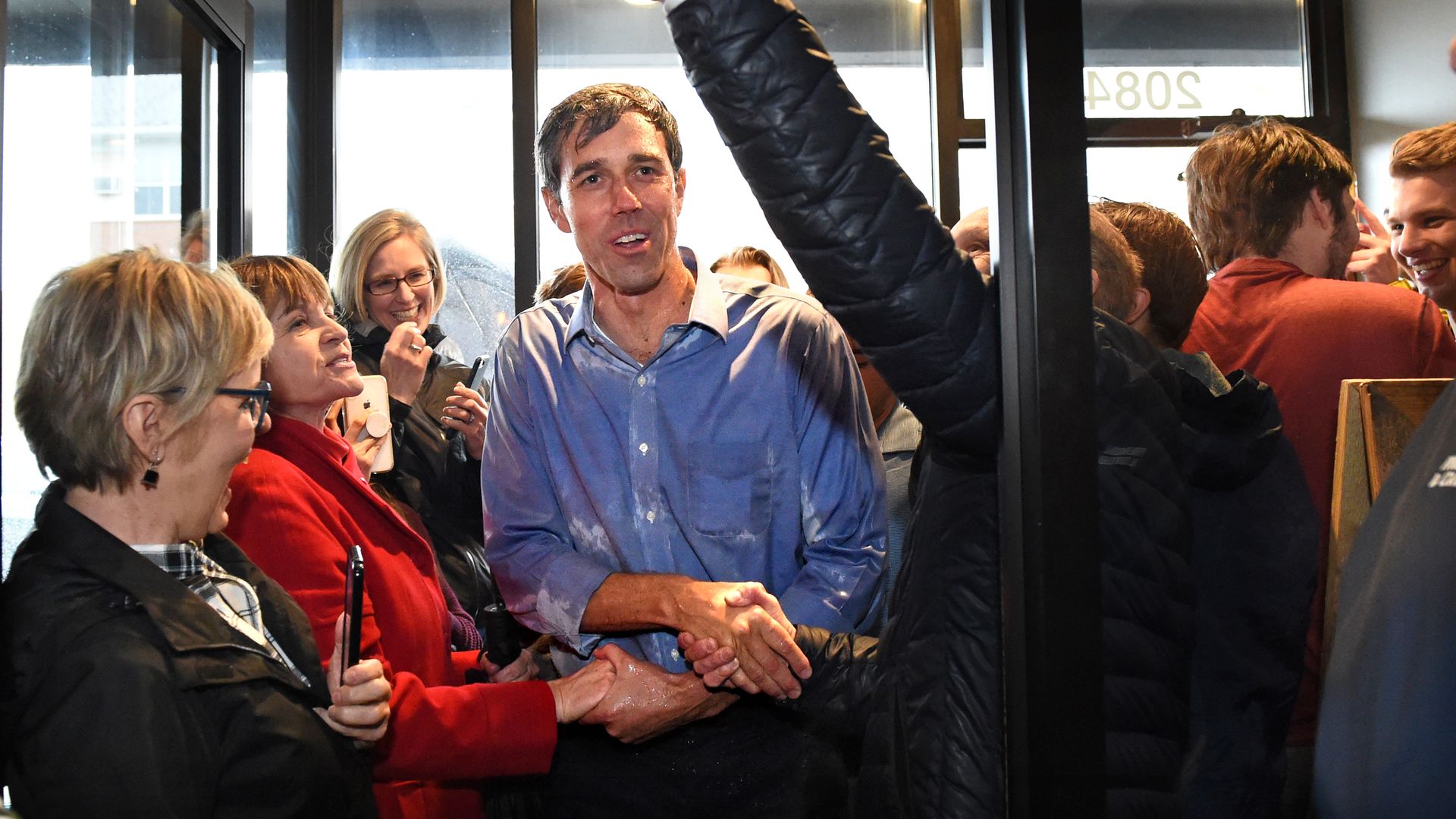 In this image, Beto stands soaked in rain and shaking multiple hands while walking through a door.