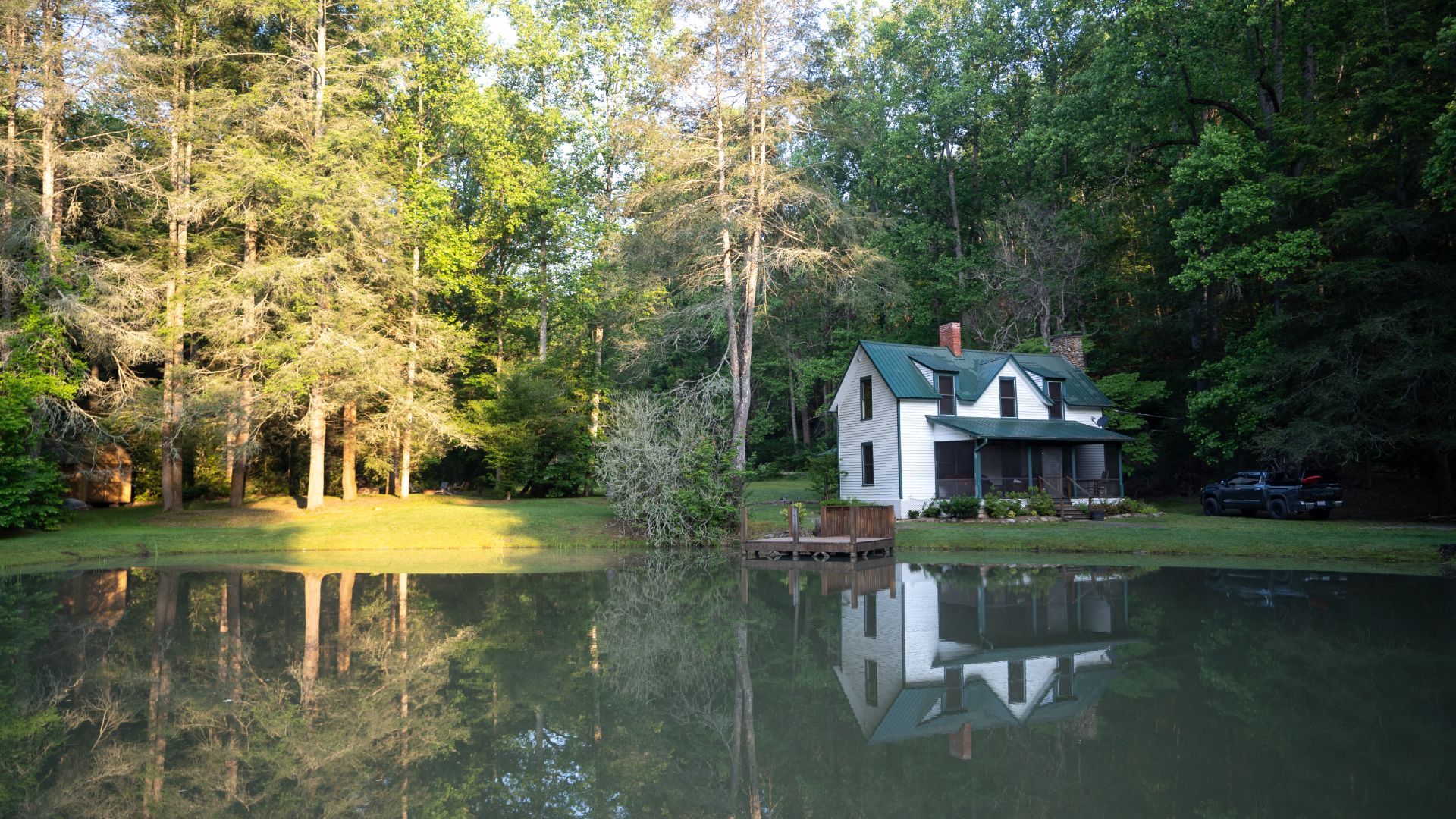 A white cabin with a green roof sits by a still pond, surrounded by tall trees. 