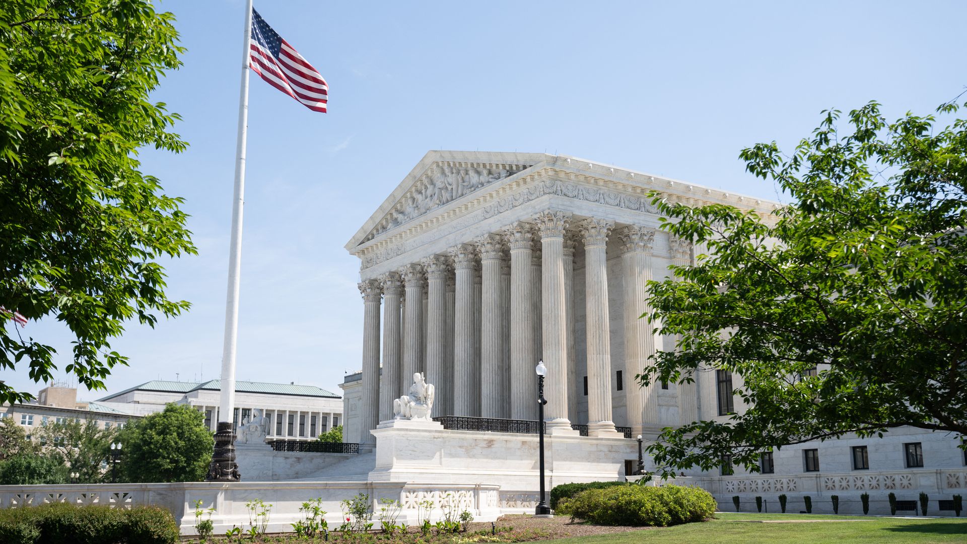 White marble Supreme Court building with tall columns, a waving American flag on a flagpole, green trees on both sides, a statue by the steps, under a clear blue sky.