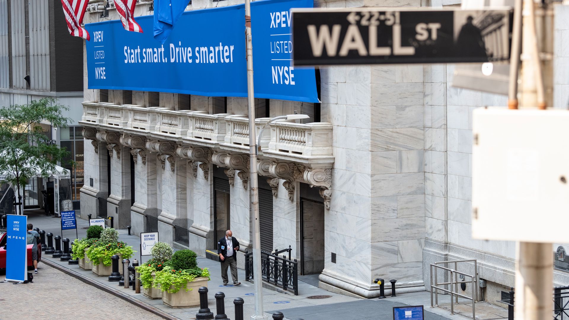 A sign for "Wall Street" in New York City, with a trader wearing a mask leaving the New York Stock Exchange in the distance