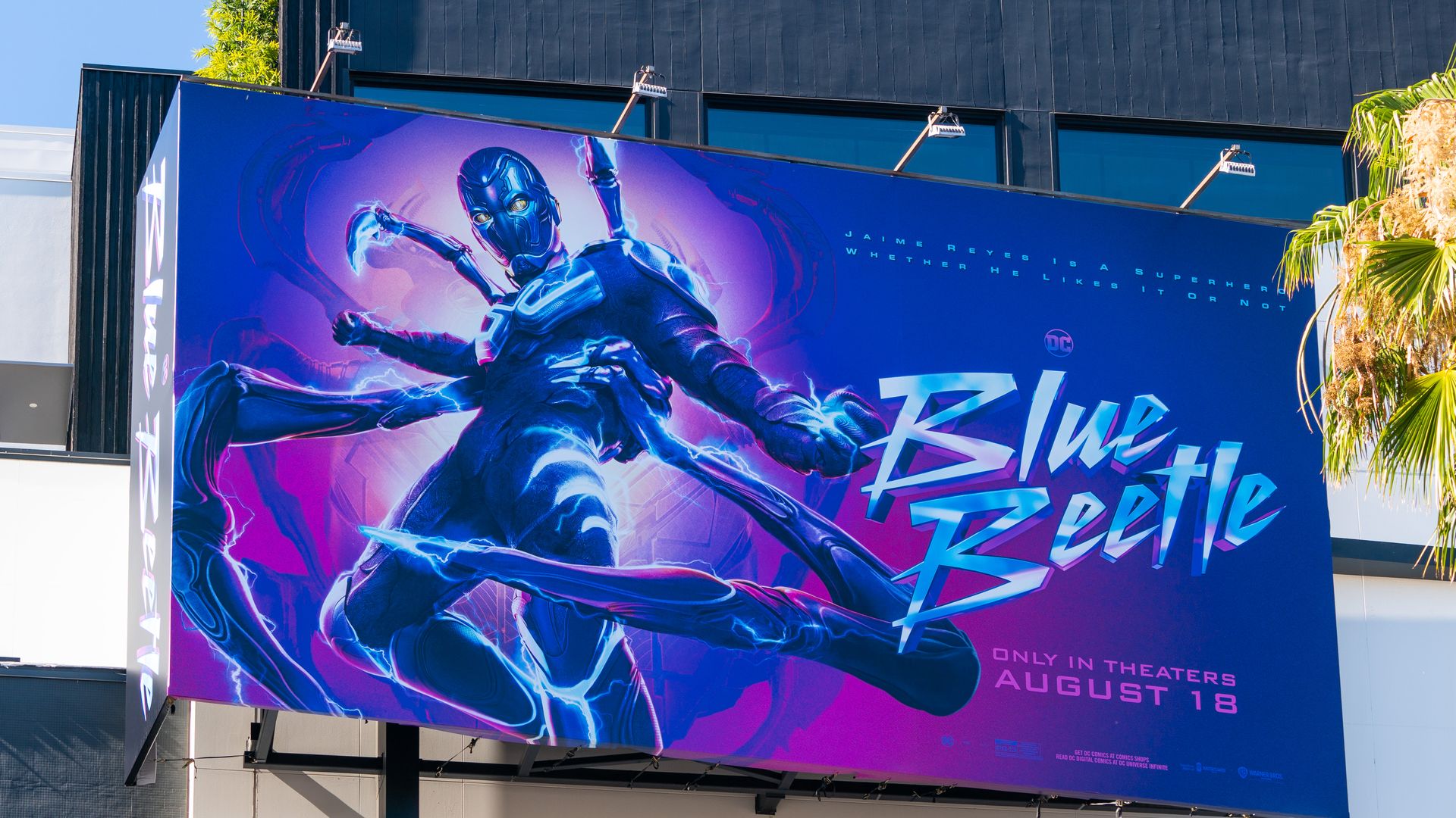 A billboard promotes the release of Blue Beetle, written in electric blue font beside the super hero. 