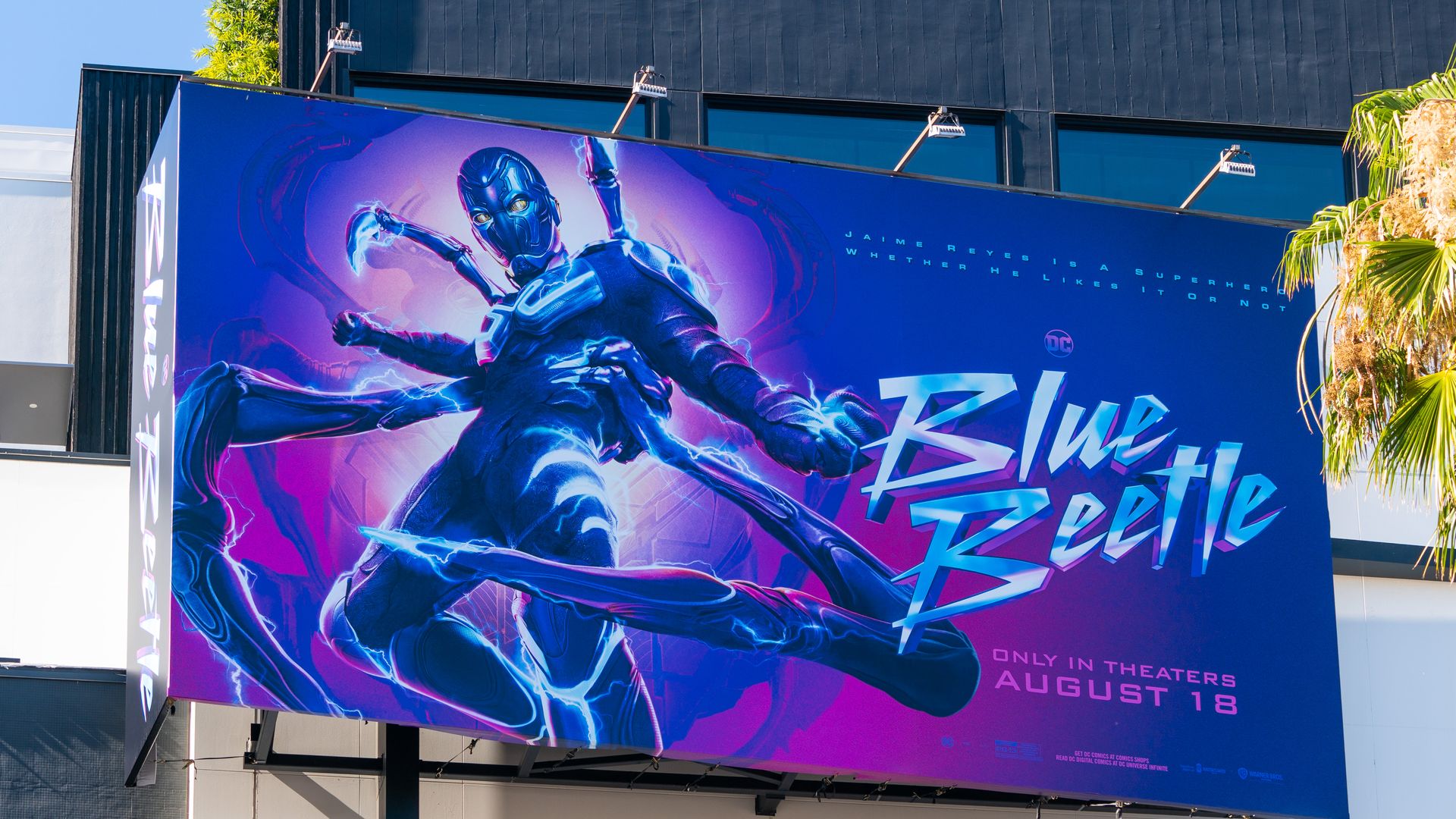 A billboard promotes the release of Blue Beetle, written in electric blue font beside the super hero.