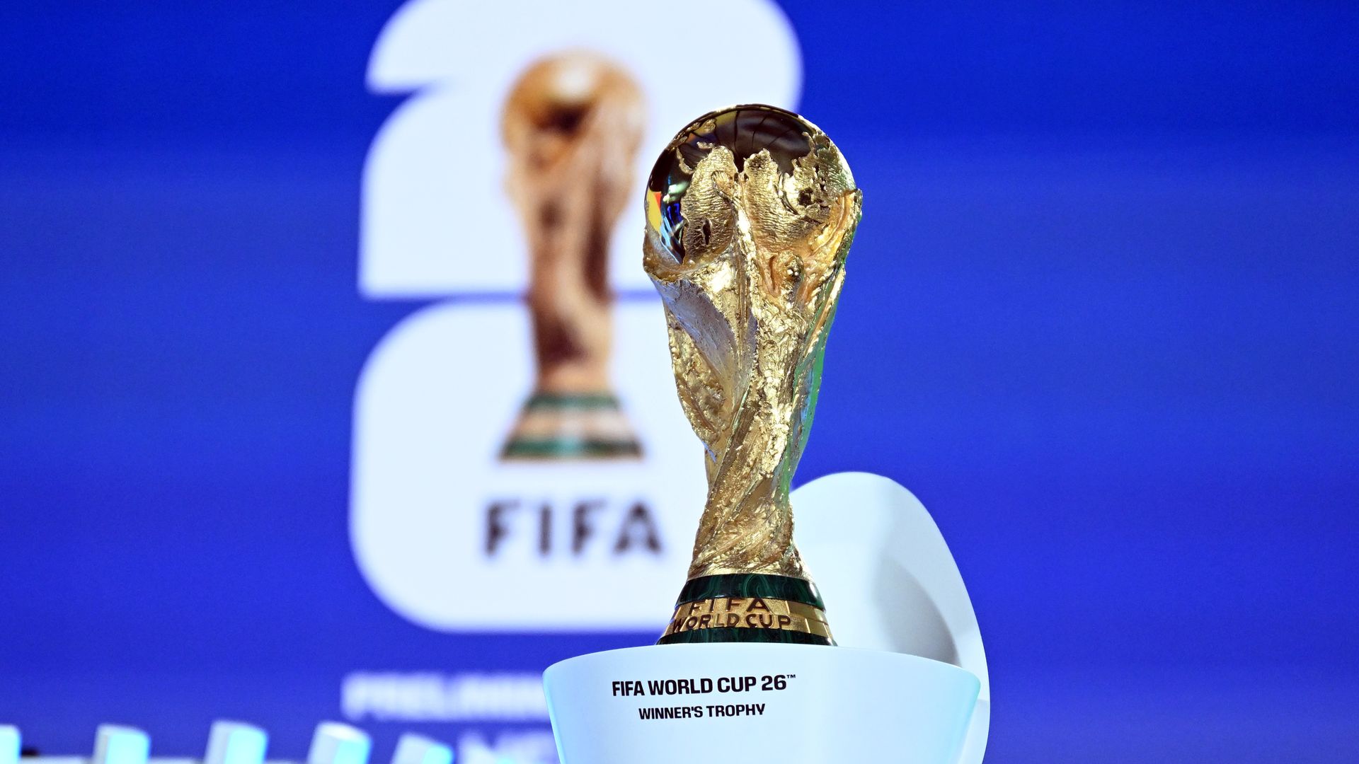 Golden FIFA World Cup 26™ Winner's Trophy on a white pedestal with blue background and blurred FIFA logo behind it.