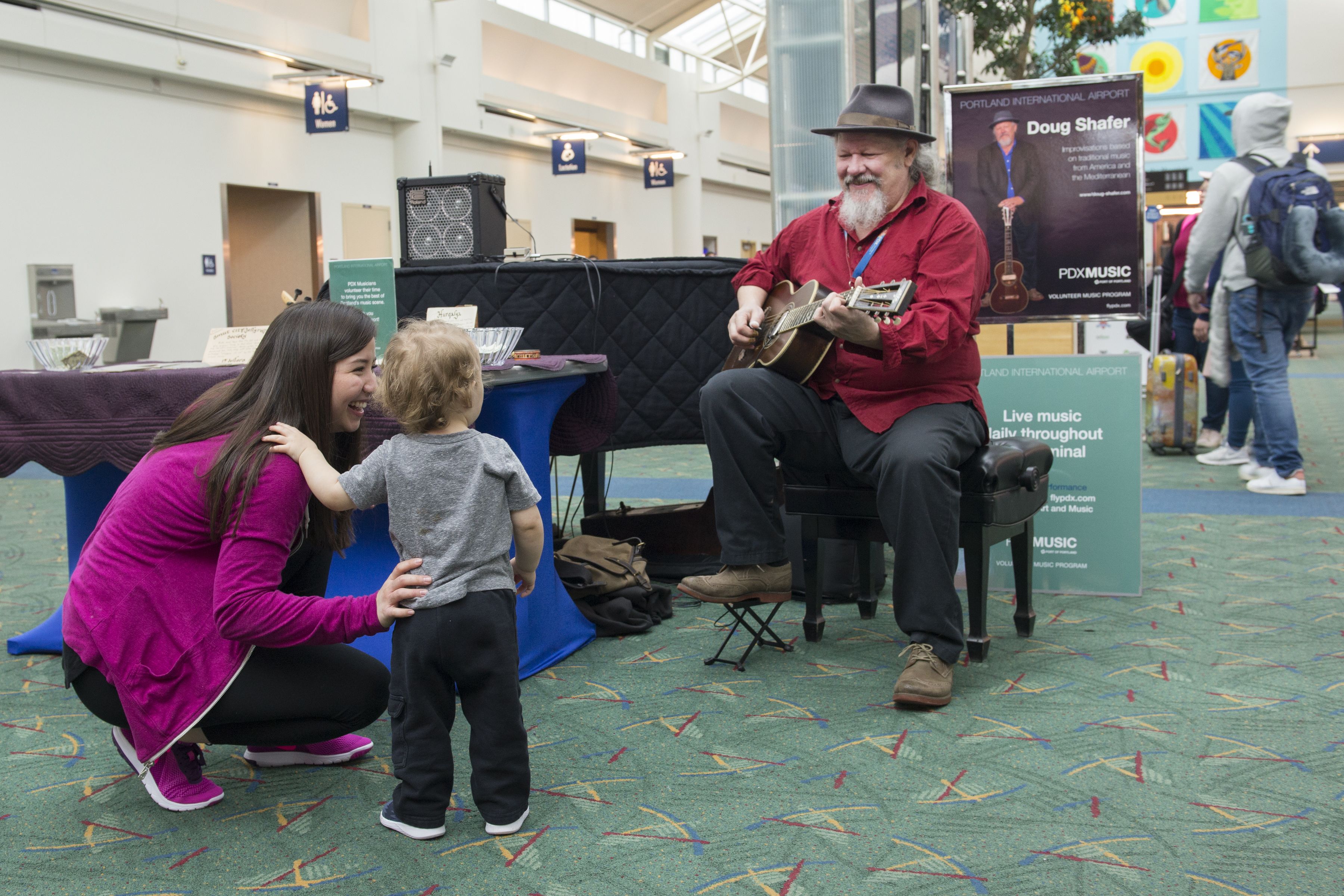Man in red shirt and hat playing guitar at airport, smiling at a toddler and woman in pink jacket who watch and enjoy the music near a PDX Music volunteer program sign.