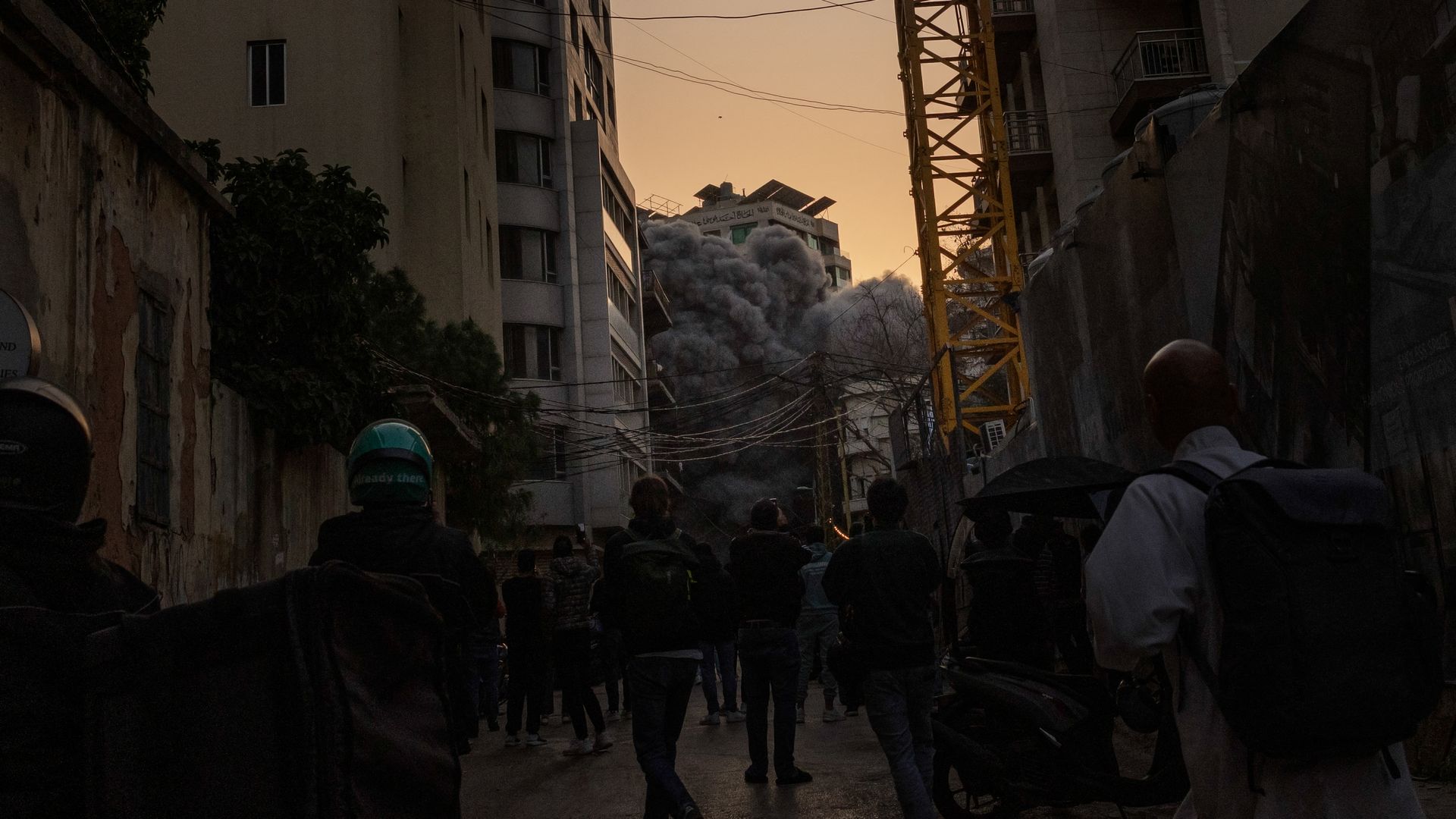 Thick gray smoke billows from a damaged building in a dense Beirut neighborhood at dusk as people gather in the street watching.