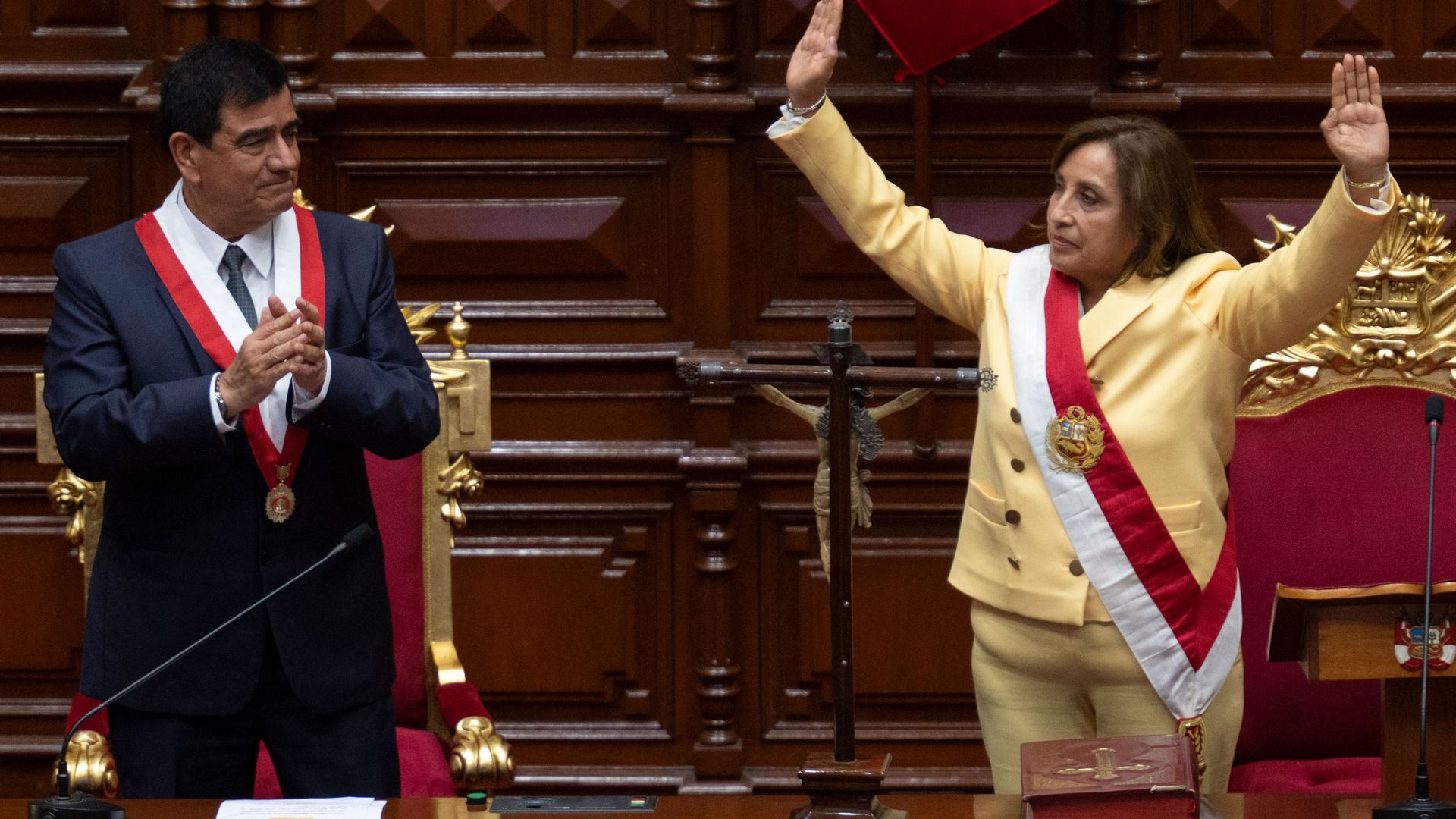 Peruvian President Dina Boluarte, in a yellow suit and a sash across her chest, raises both arms up in the air as she's sworn in 