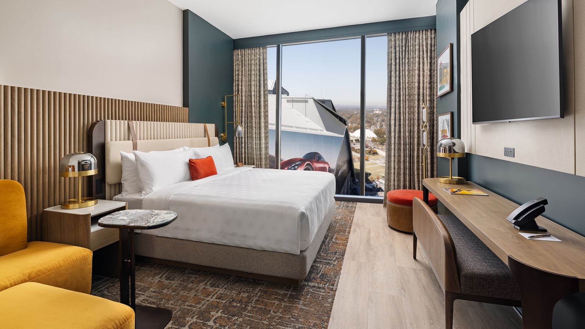 Modern hotel room with king bed featuring white linens and orange pillow, mustard yellow chair, wooden desk, large window with a partial view of Mercedes-Benz Stadium, and patterned curtains.