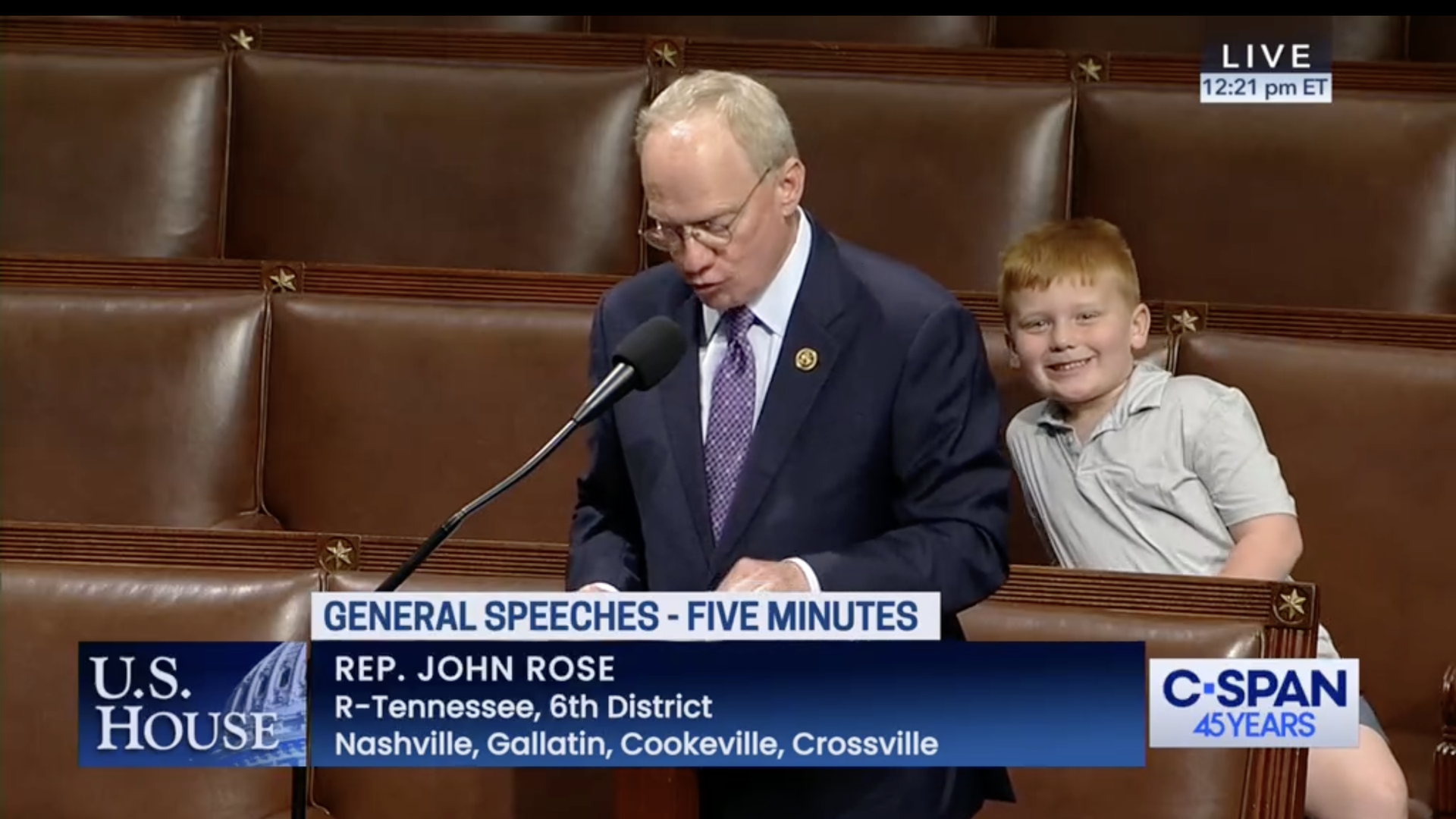 A child smiles behind a solemn speaking man.