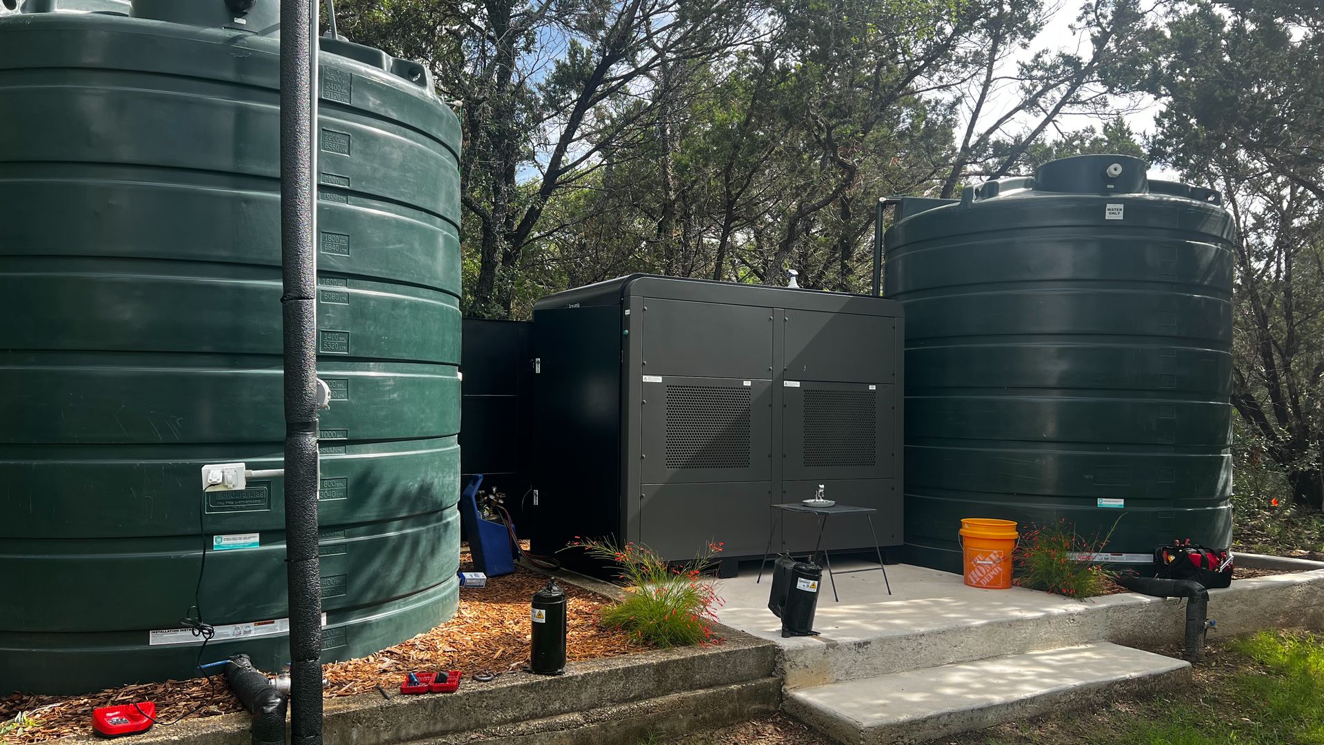 An outdoor area with a large Aquaria atmospheric water generator, which looks like a black AC unit, in between two large green water storage tanks. Trees sit in the background at the San Antonio property.