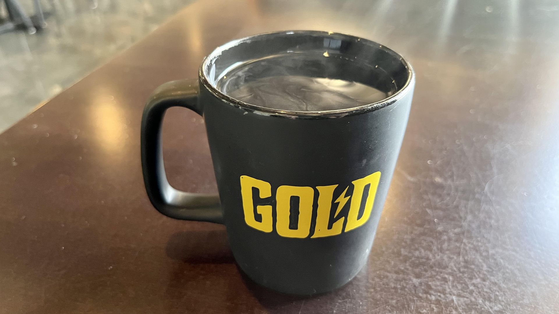 A black coffee mug with the word "Gold" in bold gold-colored text, showing steam coming off black coffee.