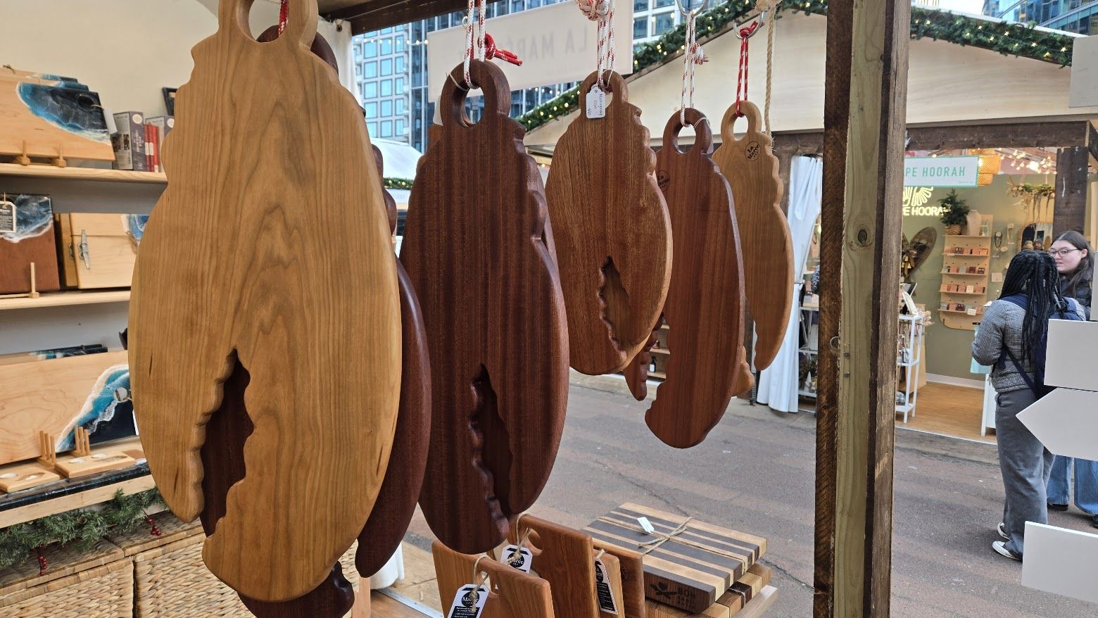 Hanging wooden cutting boards with natural edges in various shades of brown, displayed at an outdoor market stall with shoppers in the background and winter holiday decorations above.