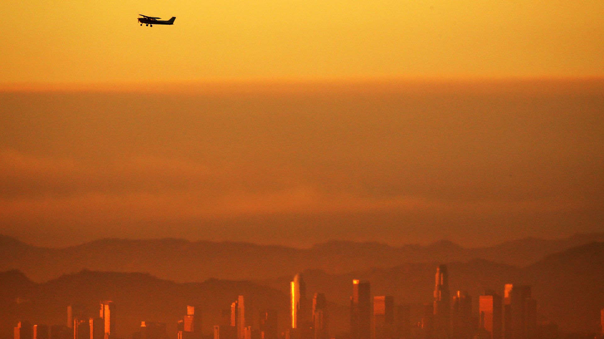 An airplane flies above an orange-yellow Los Angeles skyline. The smog nearly obscures the city skyline. 