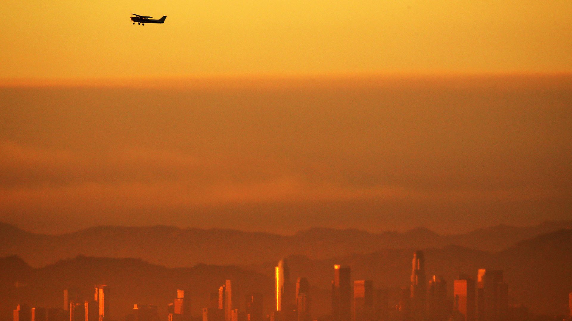 An airplane flies above an orange-yellow Los Angeles skyline. The smog nearly obscures the city skyline.