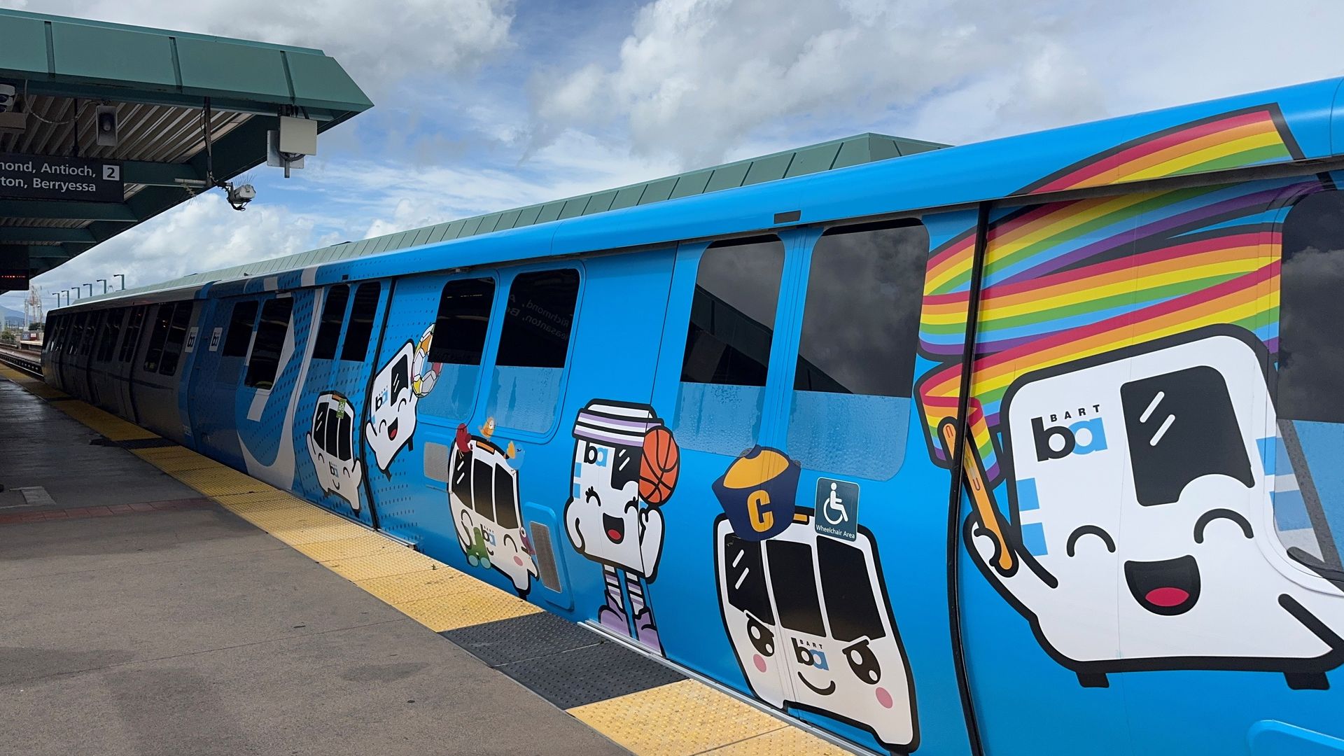 Bright blue bus with rainbow stripes and cartoon characters parked at a platform; yellow tactile paving along the edge, green station canopy above, and a cloudy sky overhead.