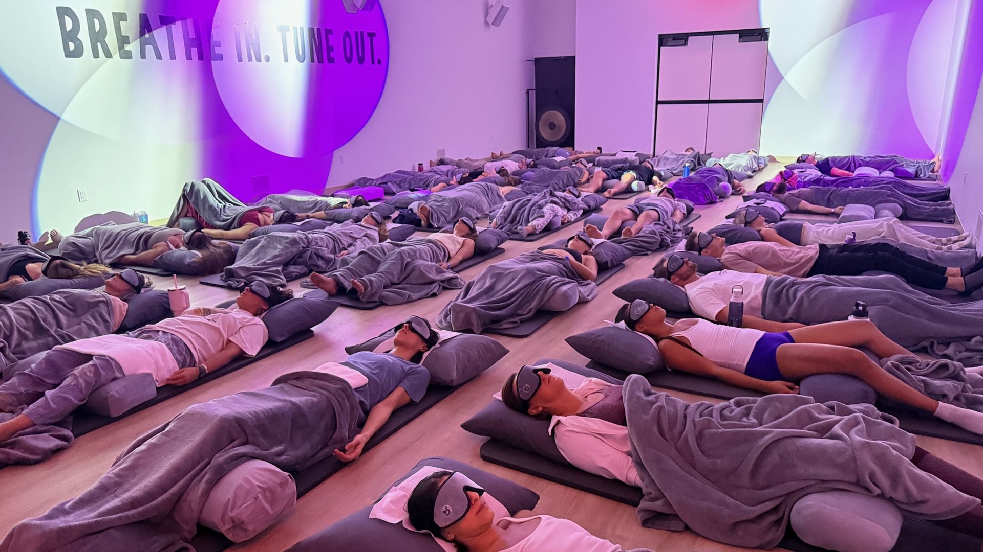 Dim, purple-lit room with dozens of people lying on mats under gray blankets, wearing sleep masks; wall reads, "BREATH IN. TUNE OUT."