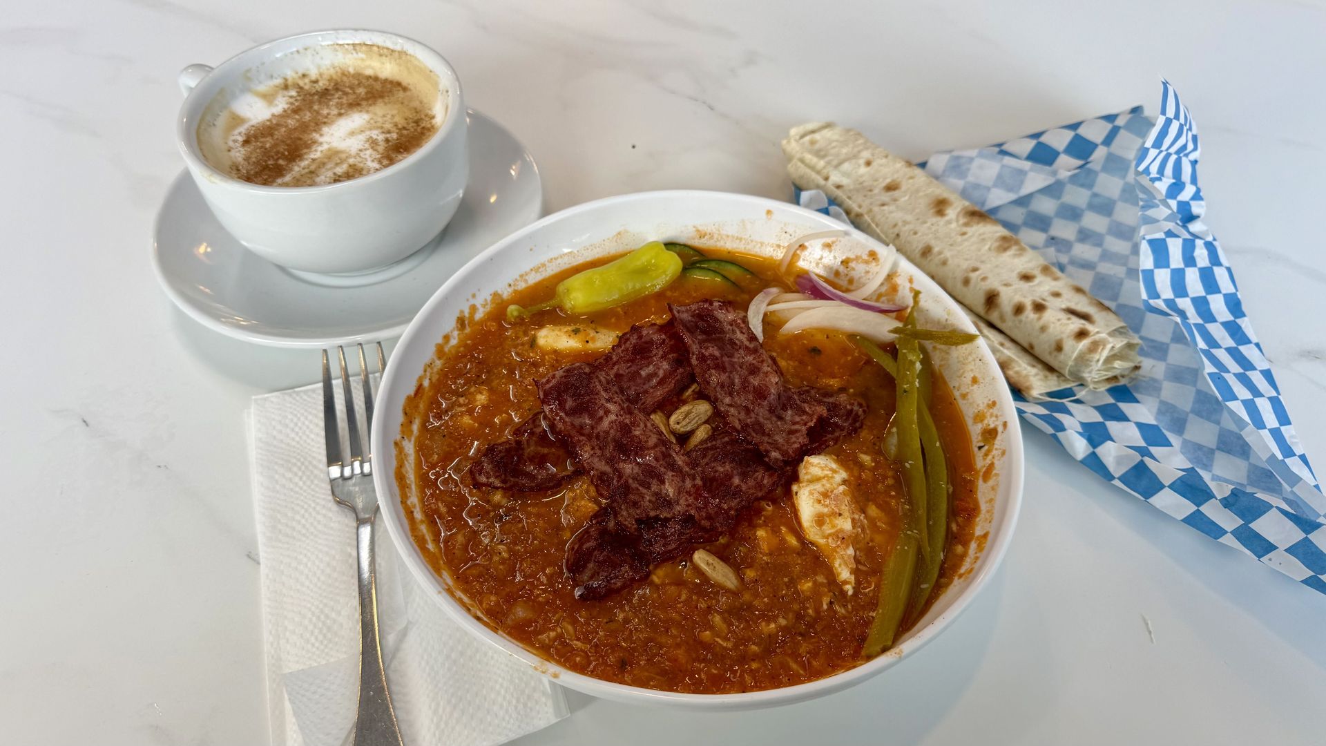 White marble table with a bowl of red stew featuring beef slices, peppers and onions, a rolled flatbread on blue-and-white checkered paper, and a cup of frothy coffee with cardamom dust.