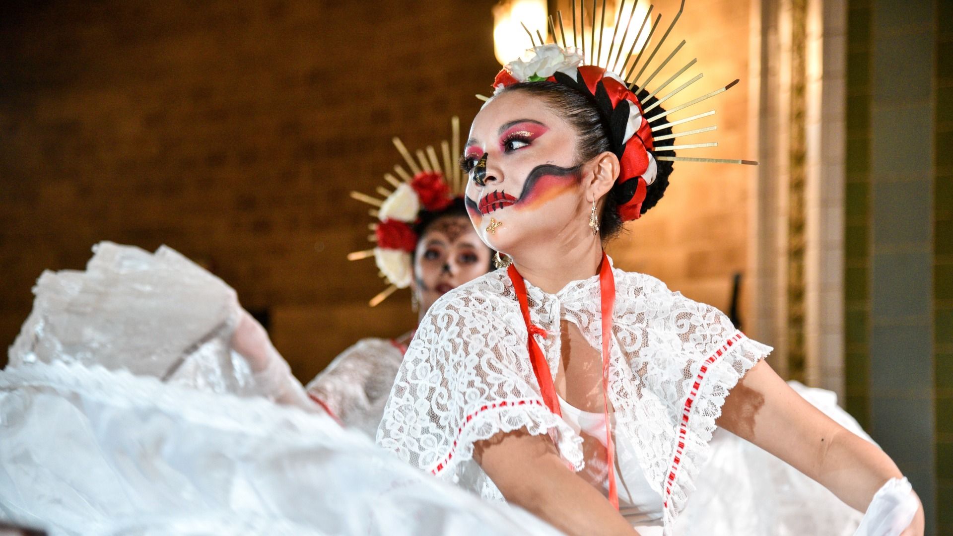 Two women in traditional white lace dresses with red accents, wearing floral headpieces and day of the dead skull face paint, performing a cultural dance indoors.