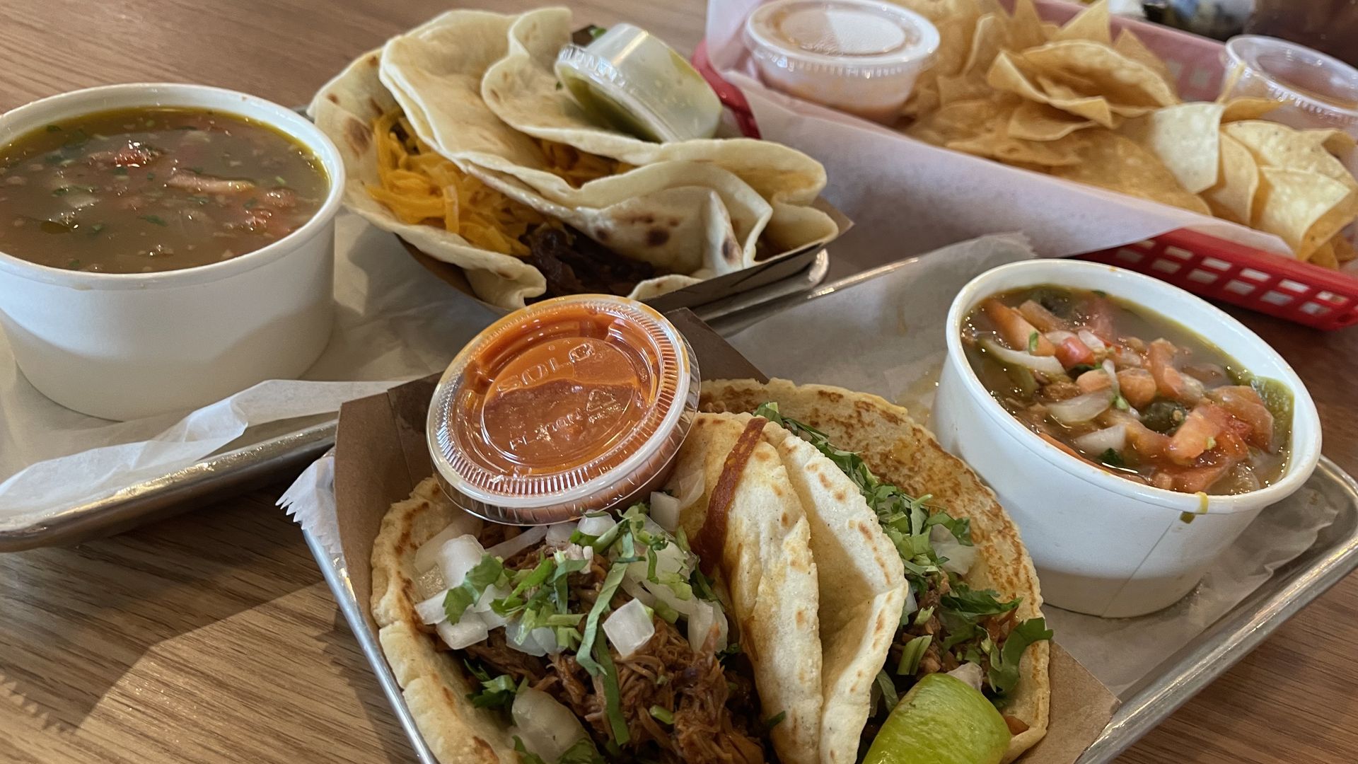 Tray with two soft tacos filled with meat, onions, cilantro, a lime wedge, and a container of red sauce, served with a bowl of soup; another tray with queso, chips, and more food in background on wooden table.