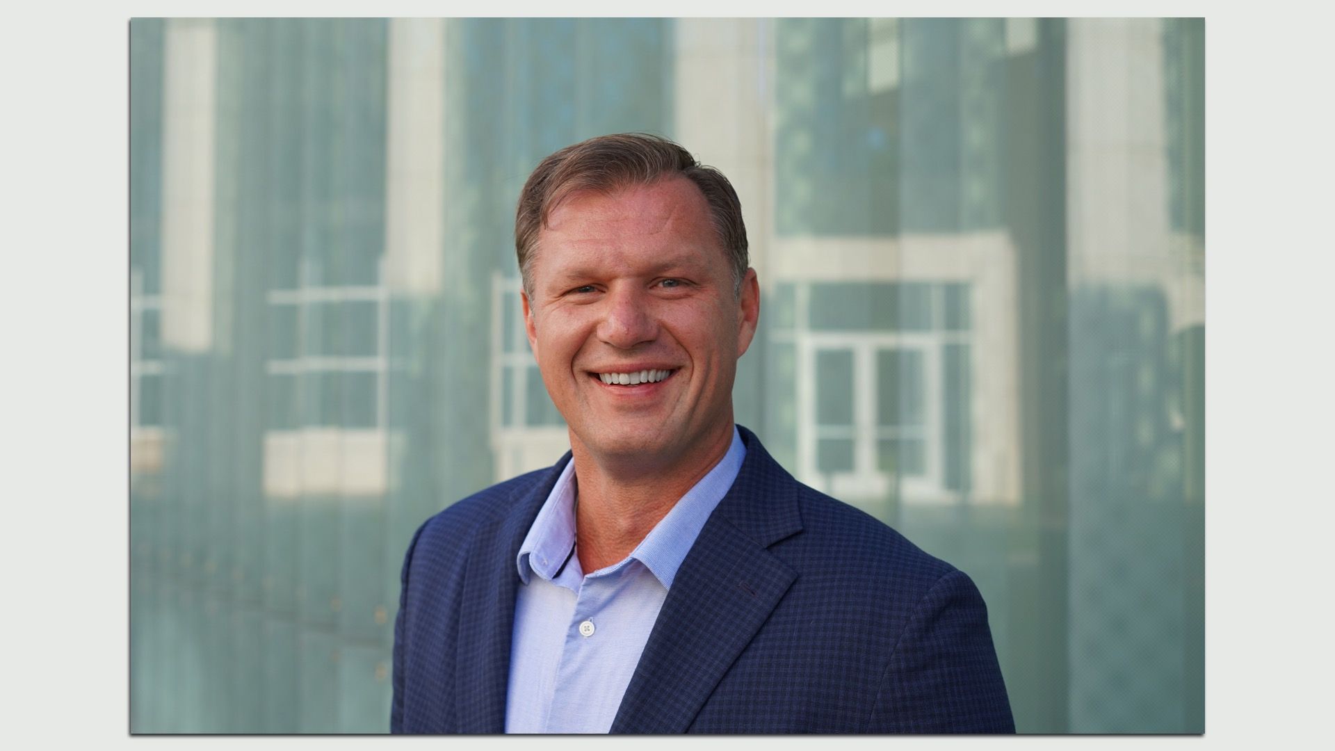 Portrait of a smiling man in a navy blue suit and light blue shirt, outdoors in front of a modern glass building.