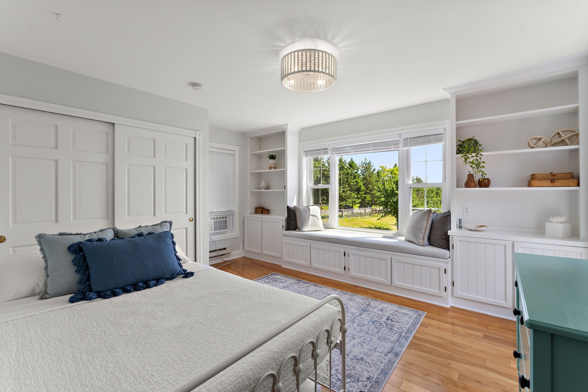 Bright bedroom with white walls, a large window seat with cushions, built-in shelves, hardwood floors, a white bed with blue pillows, and a decorative ceiling light.