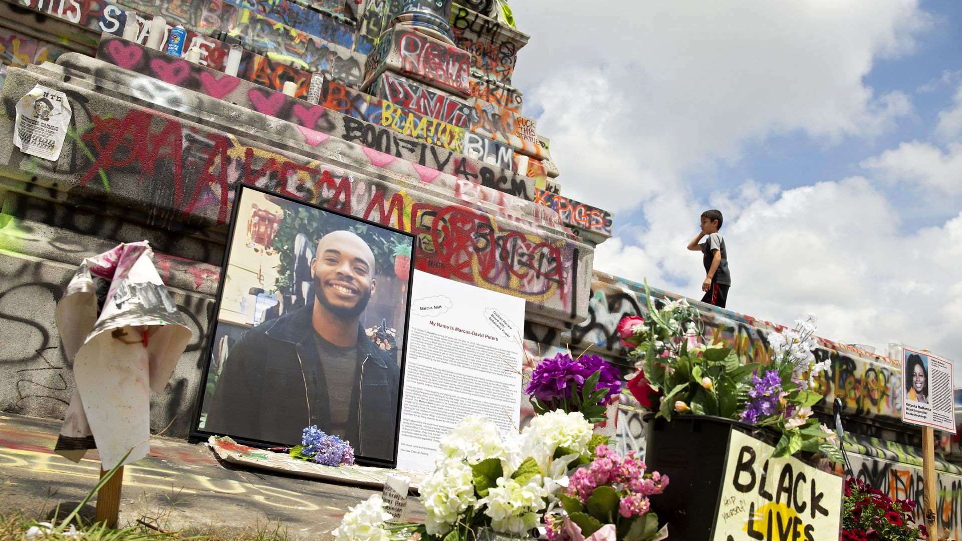 A memorial at the base of a graffitied pedestal with flowers and pictures of a man next to a sign that says "Black Lives Matter"
