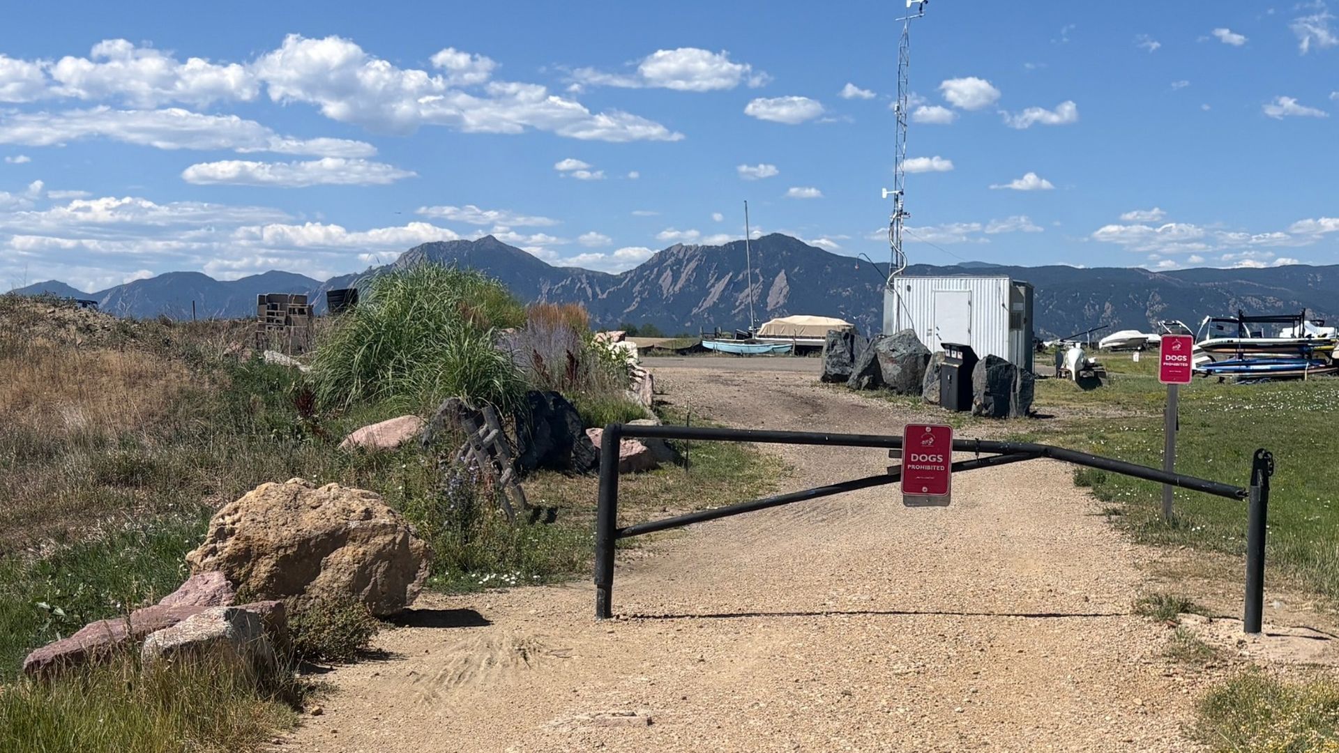 Gravel road blocked by a black gate with red "DOGS PROHIBITED" signs. A white shed, a tall antenna, and boats sit to the right; rocky foreground with grasses and distant mountains under a blue sky.