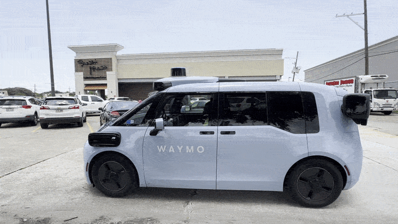 Image shows a blue Waymo SUV driving through a parking lot.