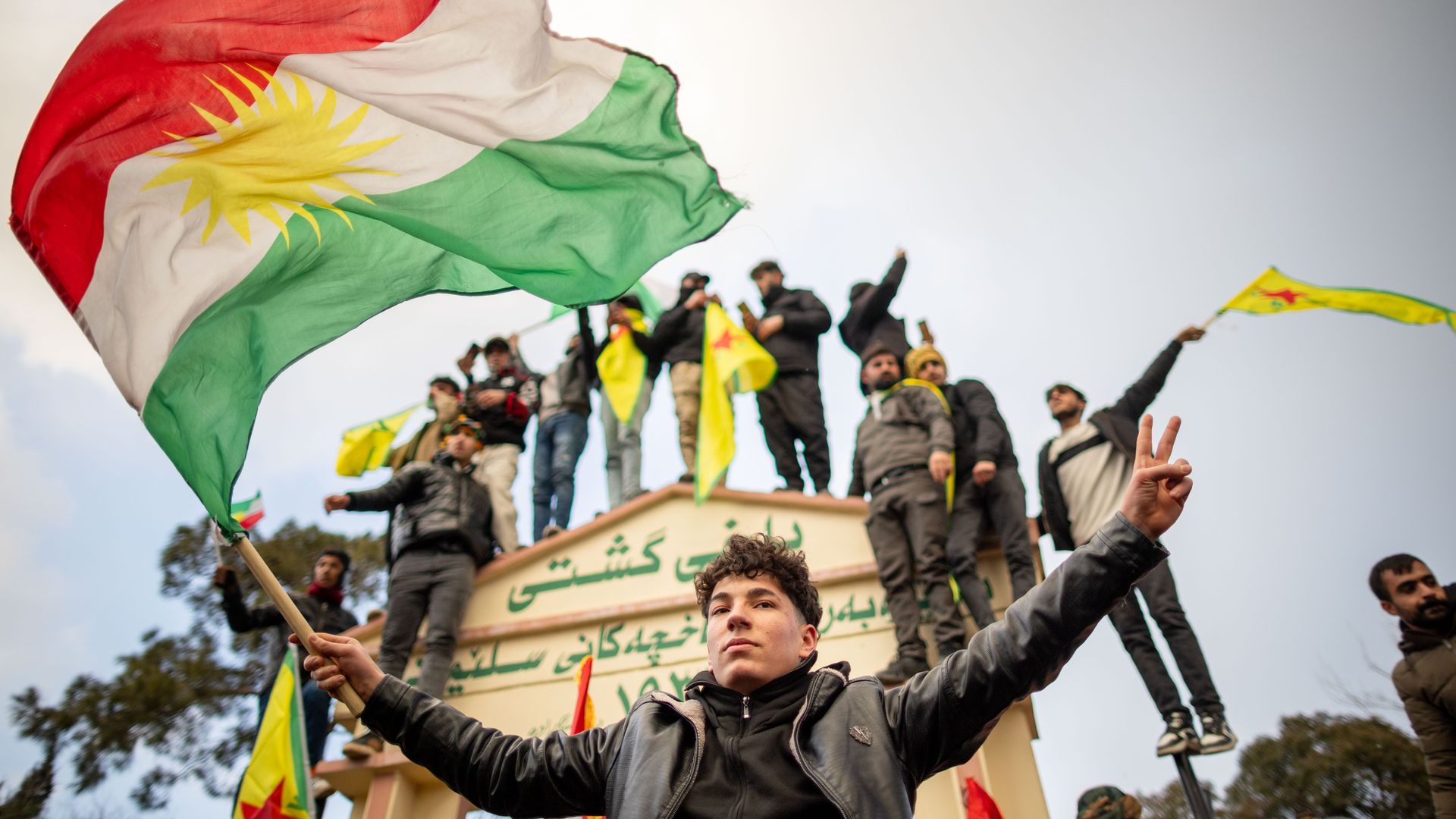 A young man holds a large Kurdish flag with red, white, green stripes and a yellow sun while others stand on a structure behind him waving yellow flags with red stars, under a clear sky.