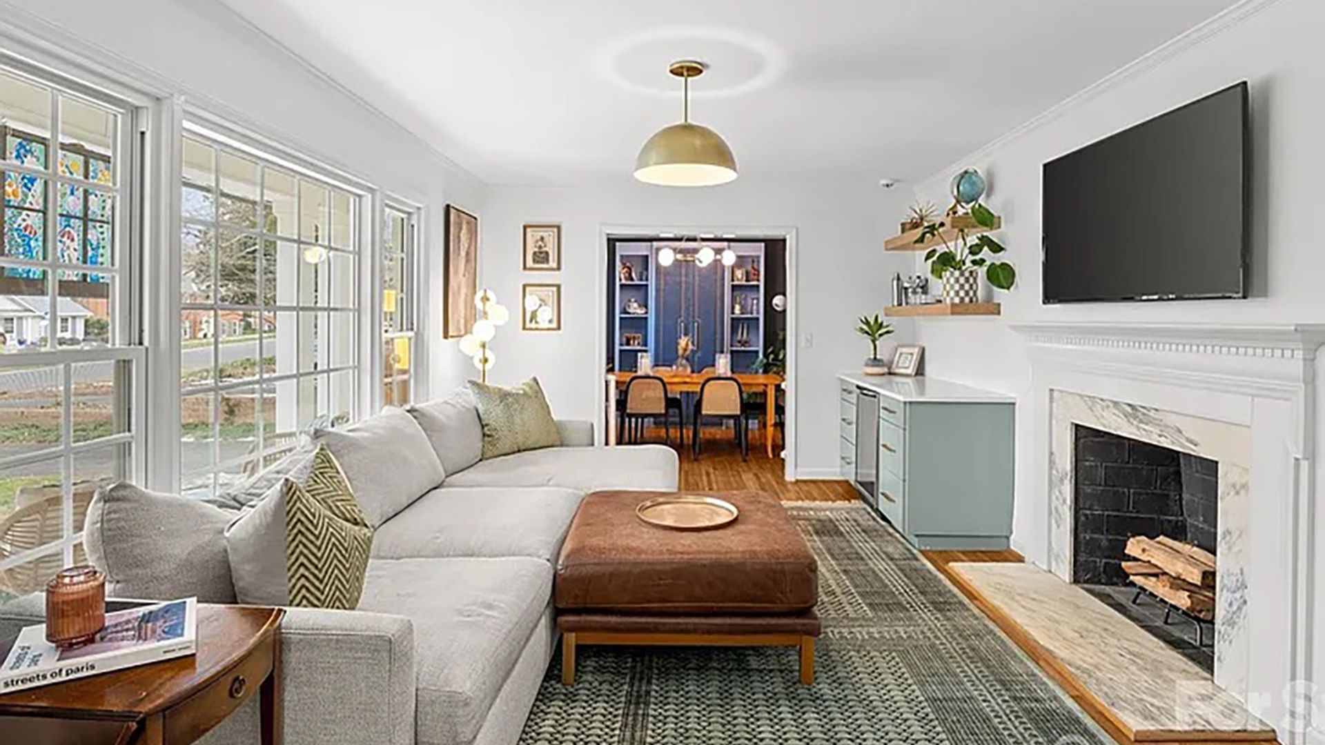 Bright living room with a gray sectional, brown ottoman, and marble fireplace with a mounted TV. Light blue cabinet, plants on shelves, large windows to the left; dining room visible through doorway.