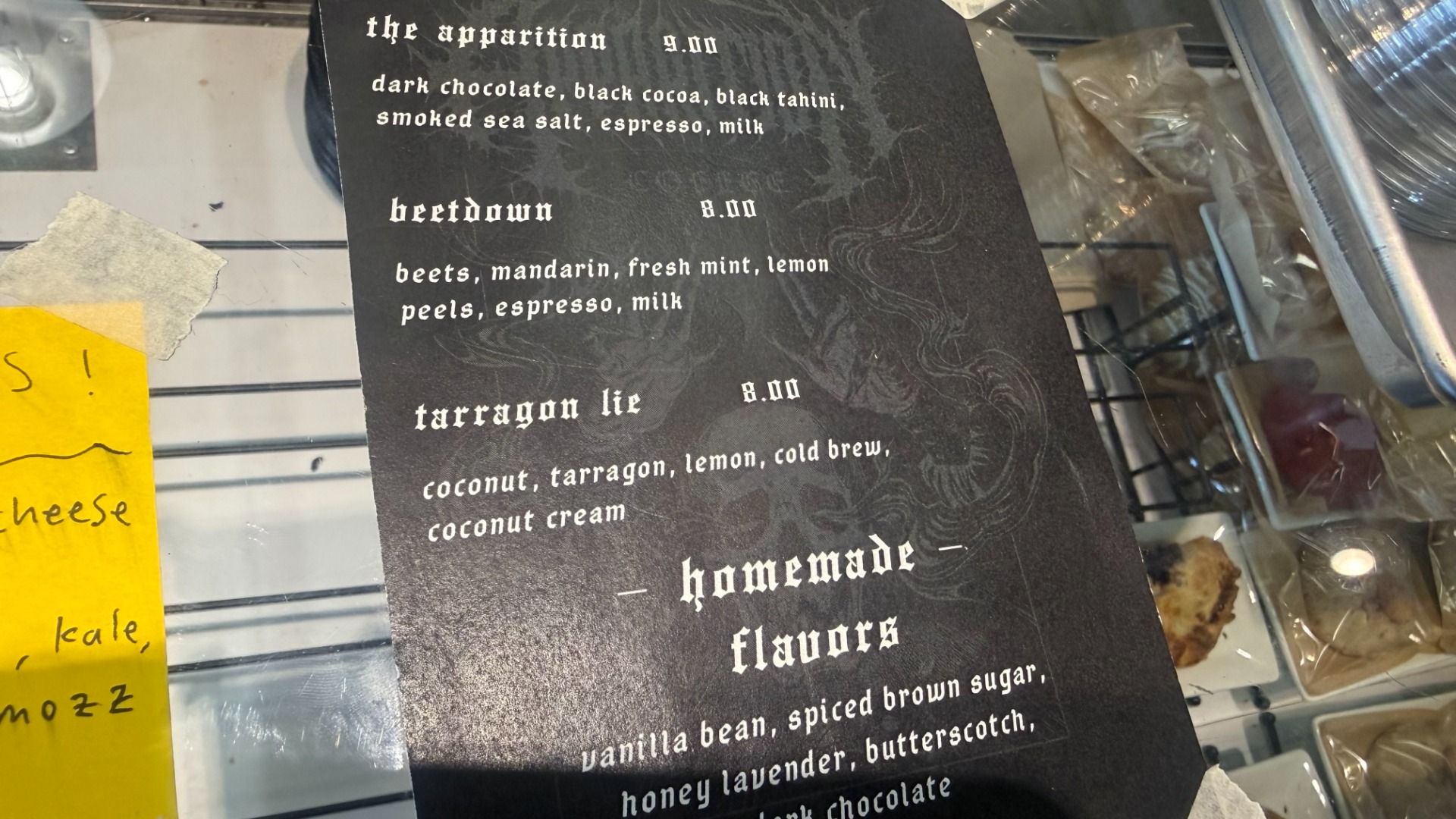Black textured menu card in a glass display with white Gothic text, listing "The Apparition" 9.00 and "beetdown" 8.00; includes ingredients like dark chocolate, espresso, milk; yellow sticky note nearby.