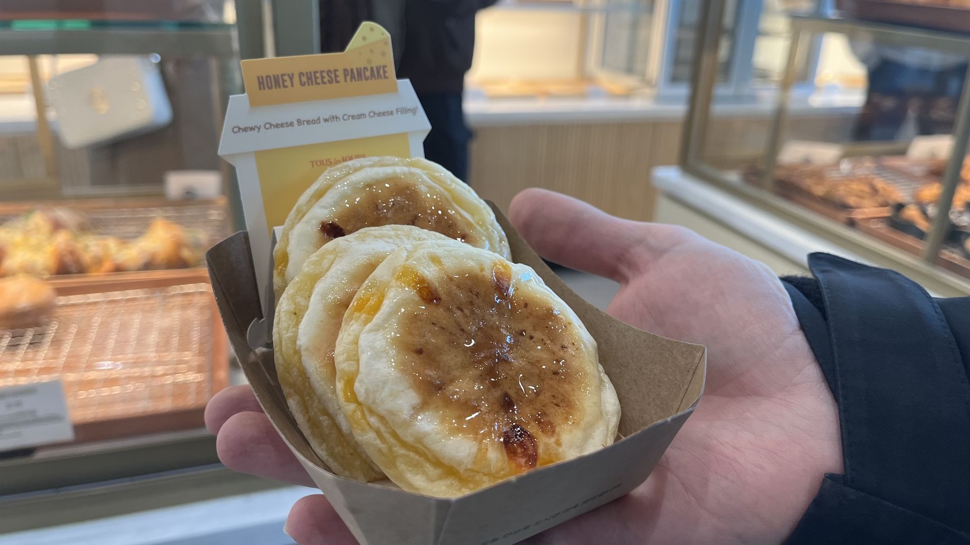 Hand holding a paper tray with two honey cheese pancakes, glazed and slightly browned on top, inside a bakery setting with blurred background.