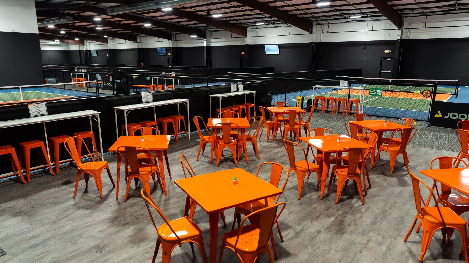 An indoor pickleball facility with orange metal tables and chairs scattered on a gray floor near indoor courts at Dill Dinkers' second San Antonio location.