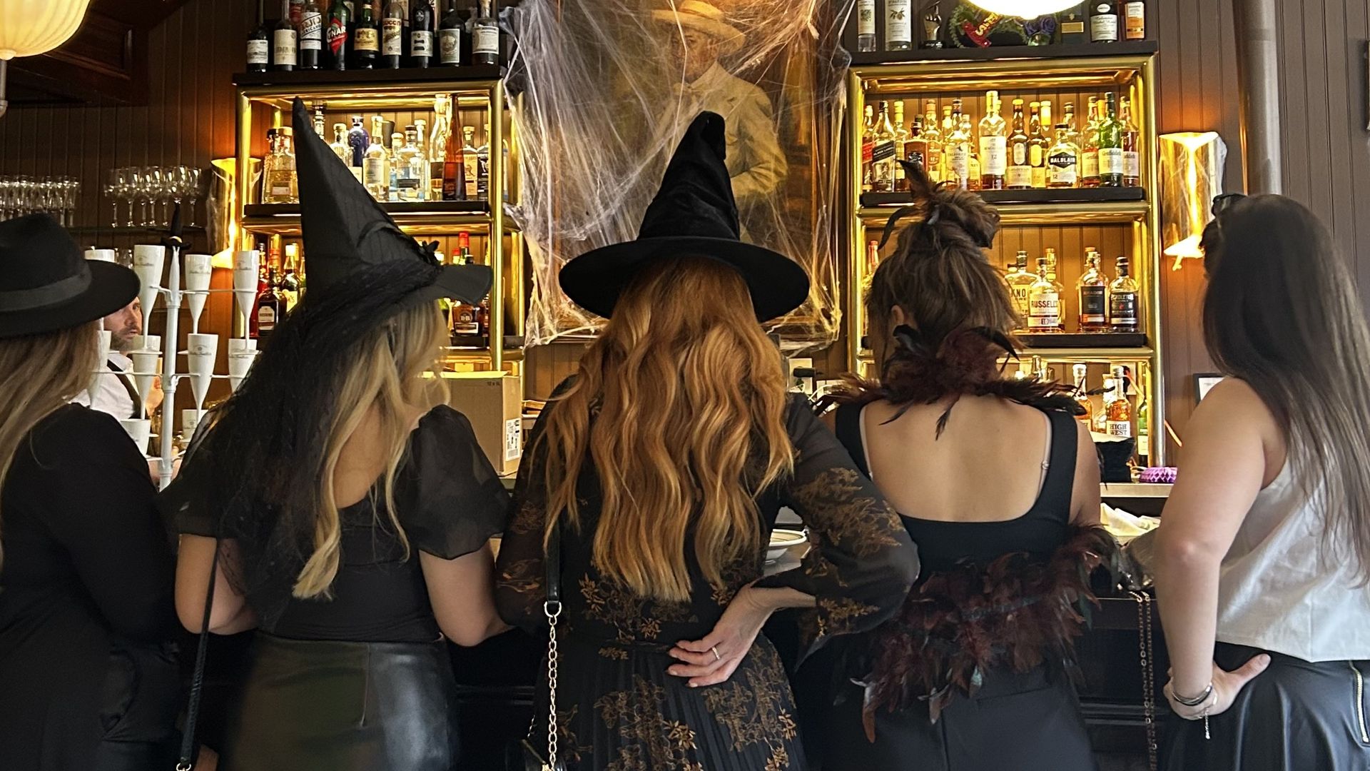 Five women dressed in black Halloween costumes, including witch hats and feathered accessories, stand at a bar with yellow lanterns and a cobweb-covered portrait behind the counter.