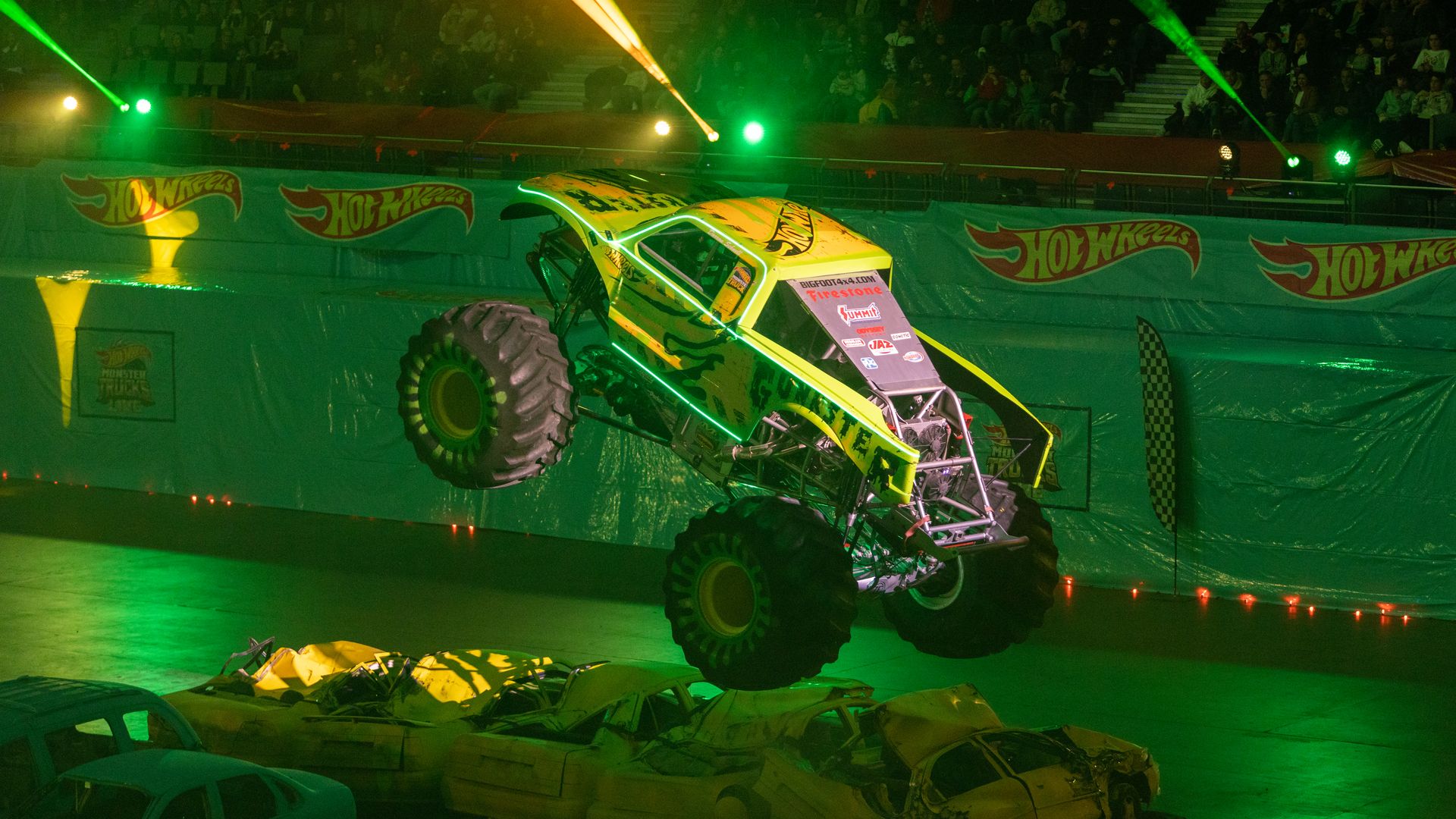 A green and yellow jumps over crushed cars in a darkened arena.