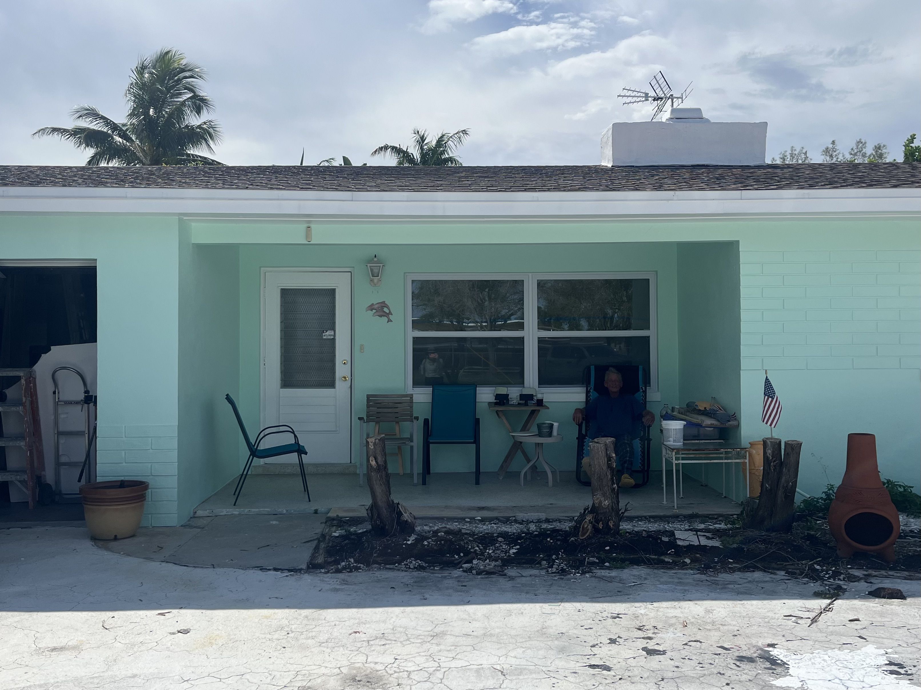 Light blue house porch with three chairs, a small table, tree stumps in front, American flag on table, orange chiminea, and a person sitting in a chair under cloudy sky with palm trees.