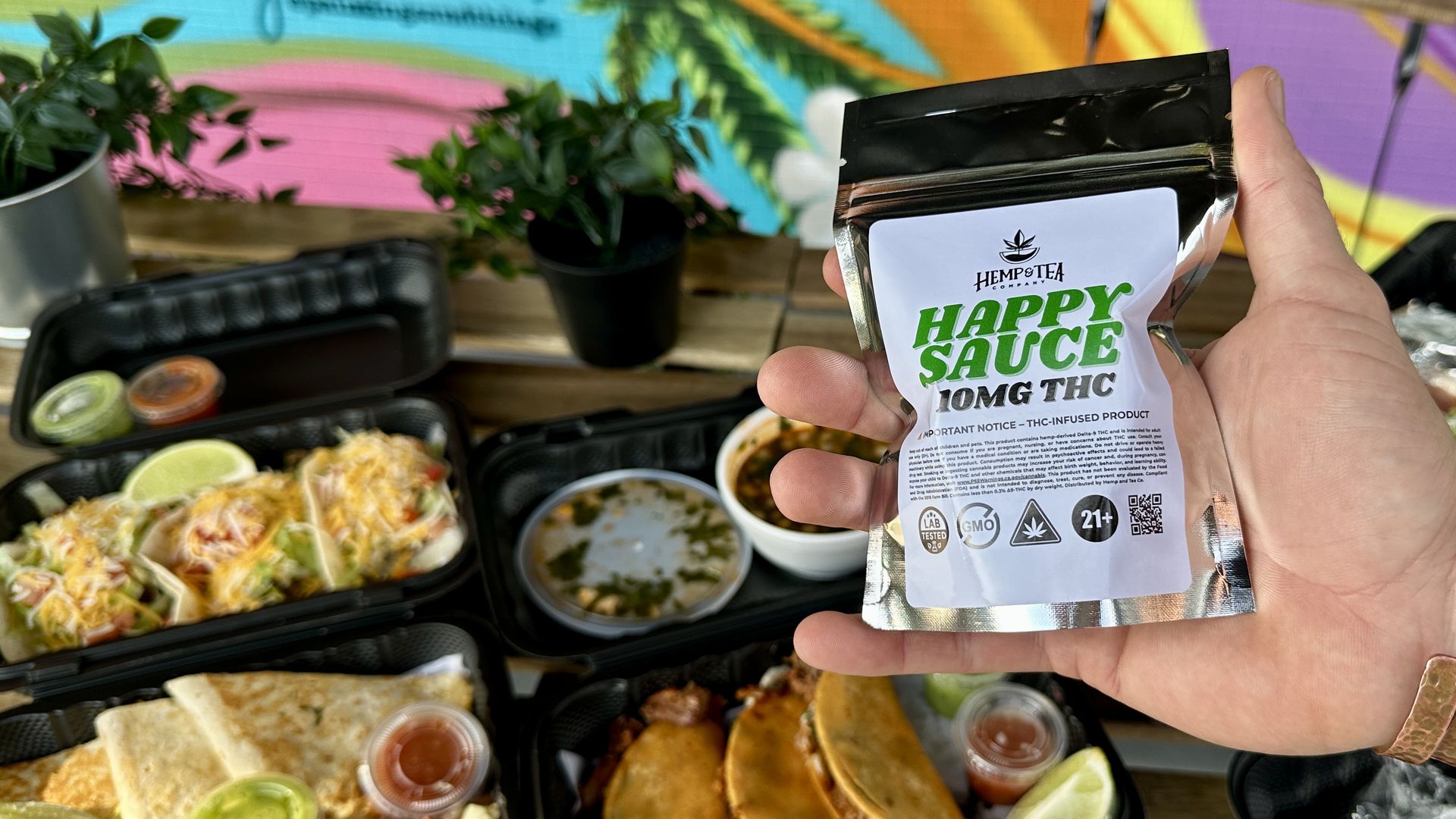 Hand holding a black and silver packet labeled "Happy Sauce 10MG THC" by Hemp Tea Company, with various Mexican food trays and a colorful mural with leaves in the background.