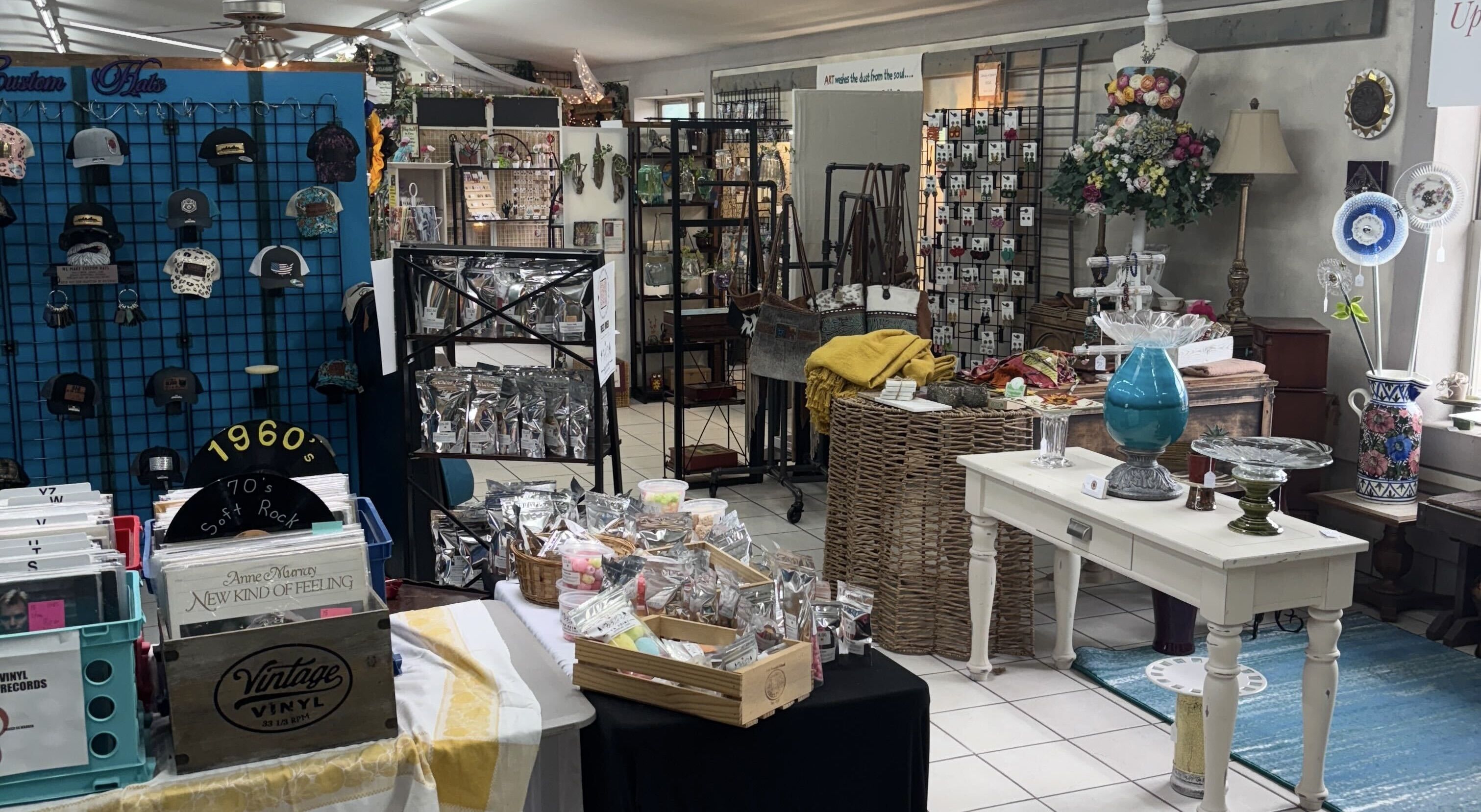 Inside a boutique with vinyl records, custom hats on a blue wall grid, bags, jewelry displays, silver packaged goods, floral arrangements, and decorative glass items on tables.