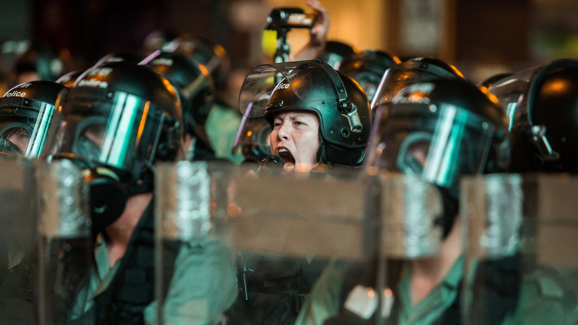 This image shows a riot officer yelling with his face exposed in a line of helmeted officers. 