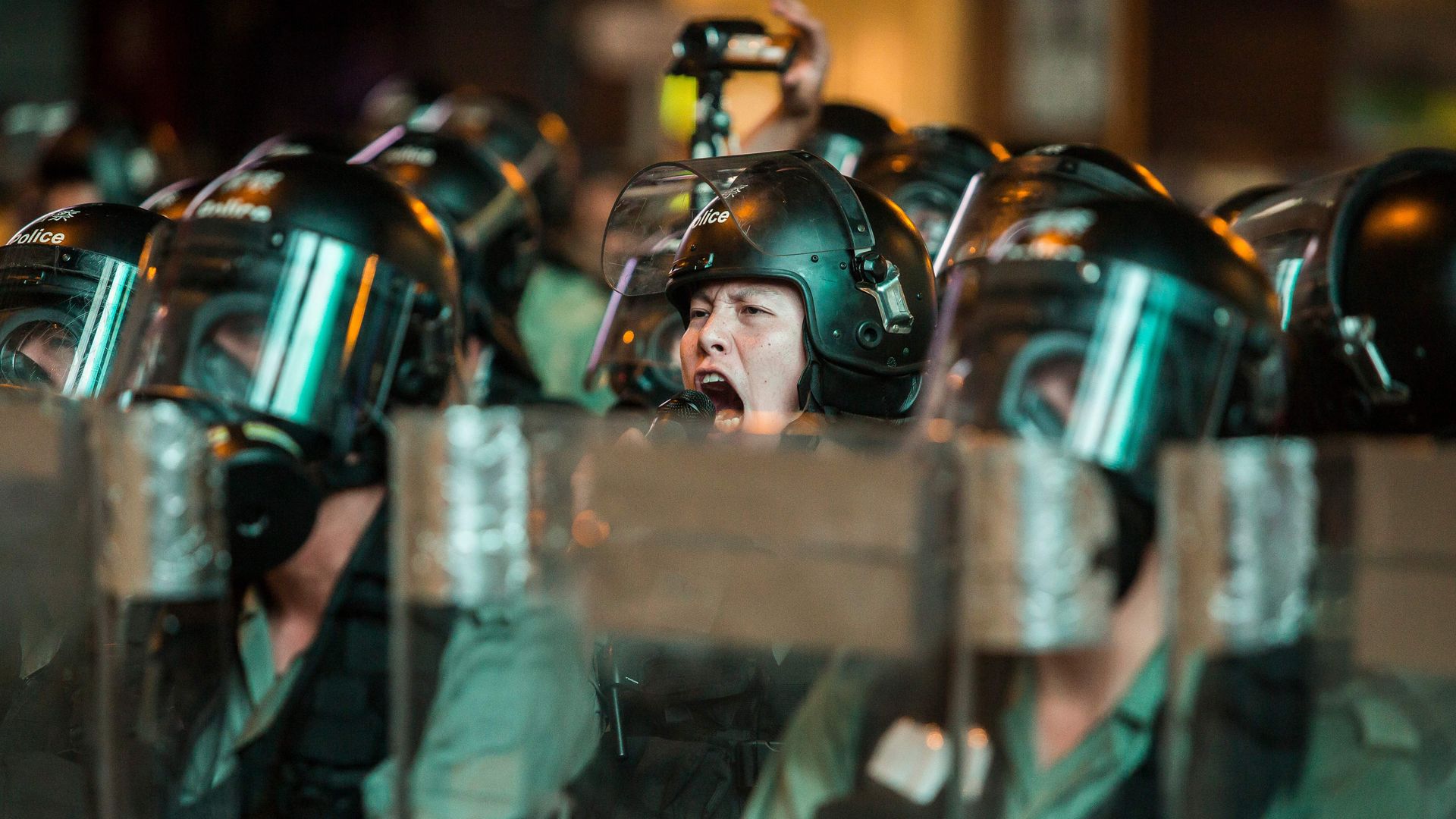 This image shows a riot officer yelling with his face exposed in a line of helmeted officers.