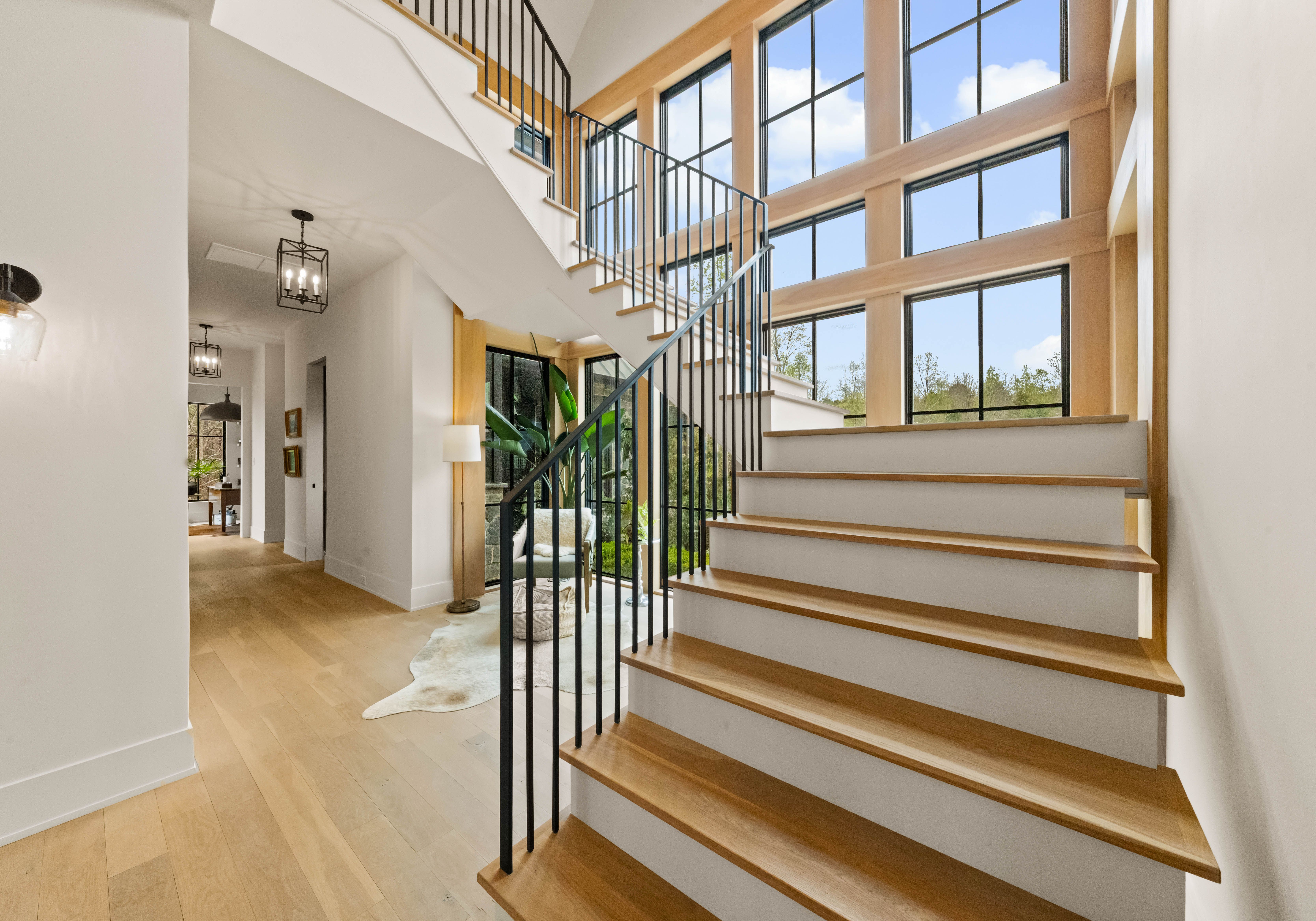 Bright, modern two-story foyer featuring a sweeping wooden staircase with black metal balusters, tall grid windows, light hardwood floors, white walls; view into a hallway with a living area and greenery outside.