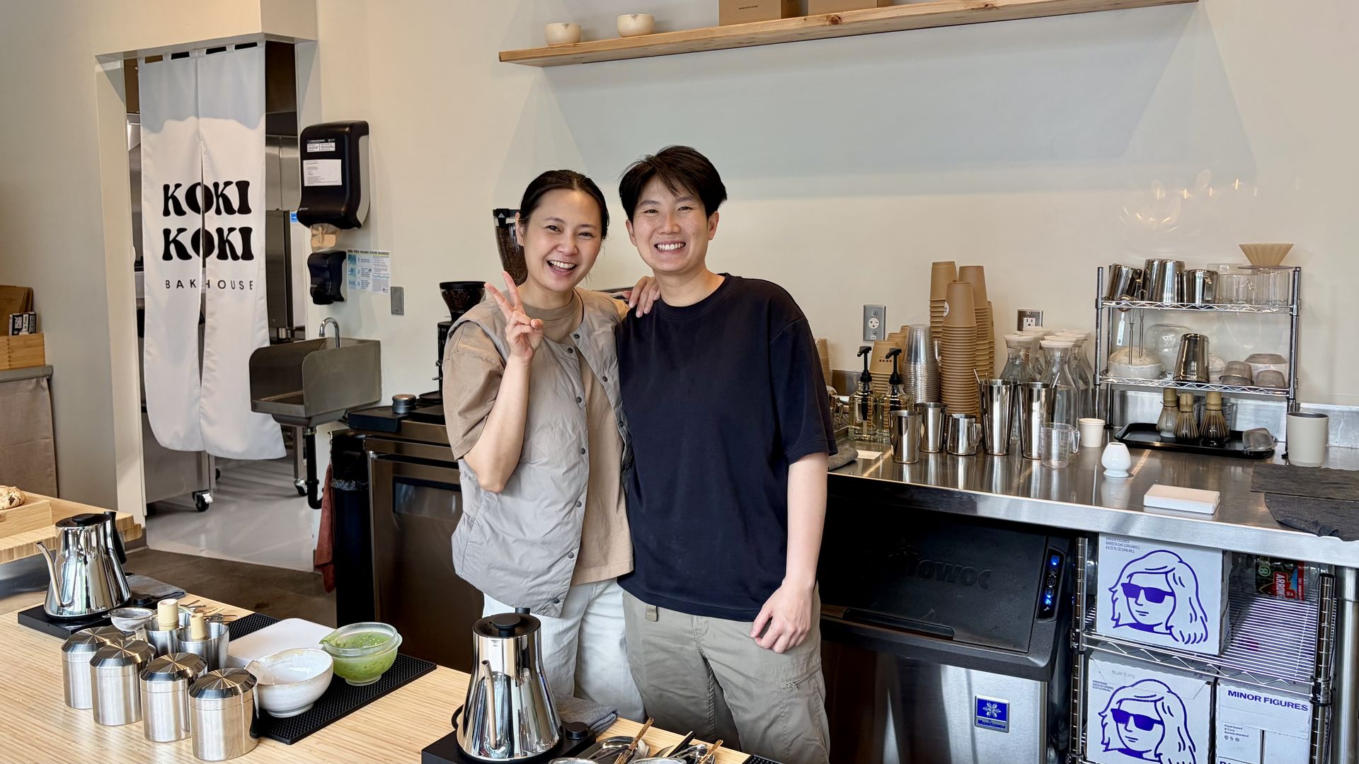 Two smiling cafe workers pose behind a light wood counter. A white curtain reads "KOKI KOKI BAKHOUSE" by the kitchen door; shelves to the right hold cups, glassware, and equipment.