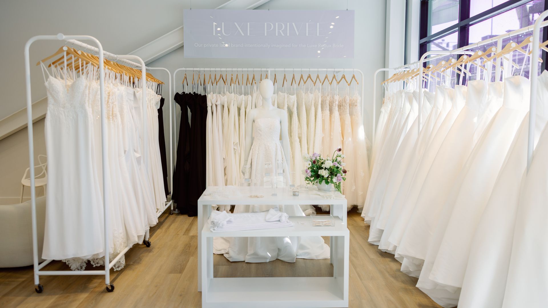 Elegant bridal boutique with rows of white wedding dresses and a few black gowns, a mannequin in a strapless white dress, and a white table with jewelry and flowers under natural light.